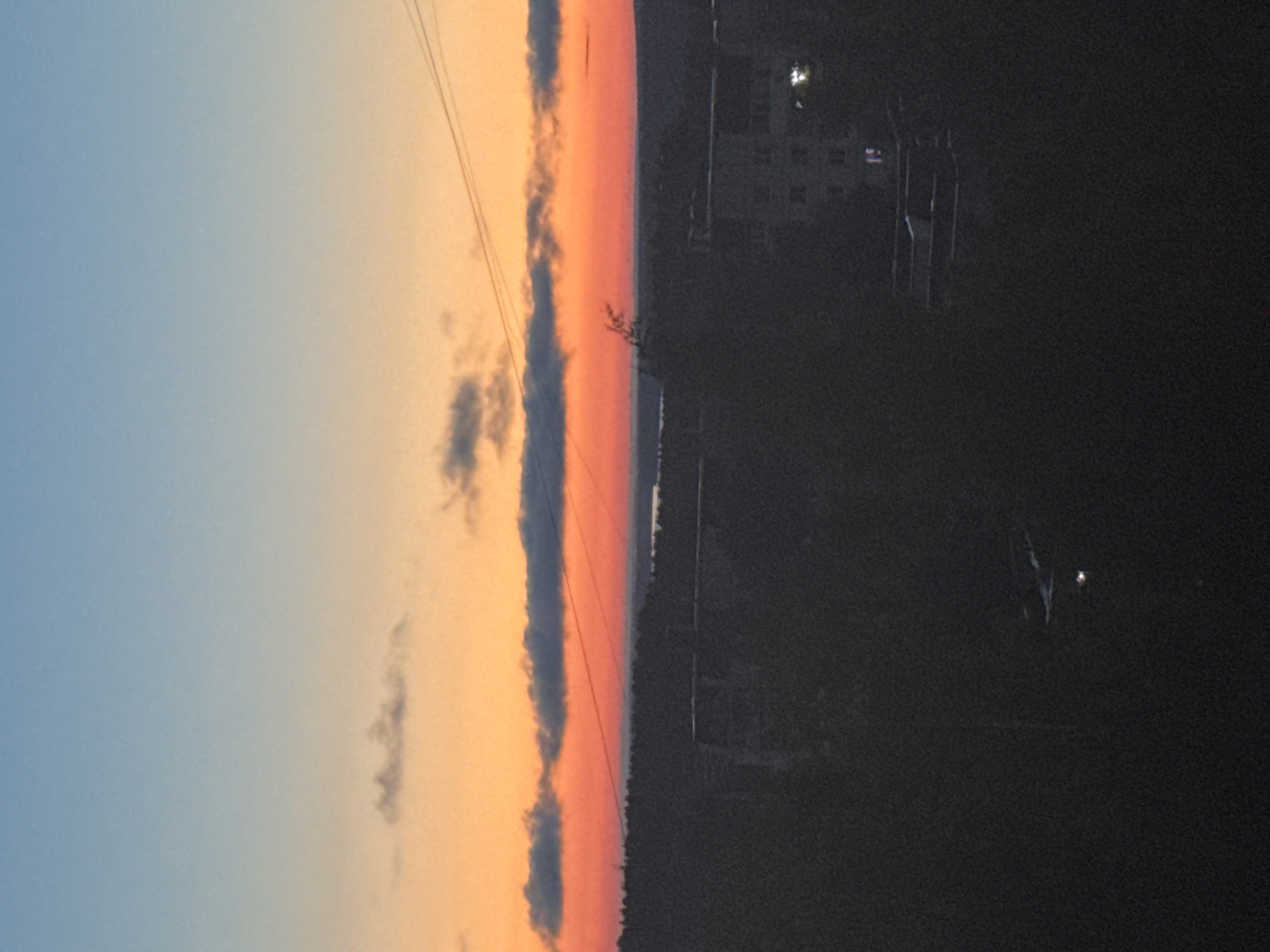 The image depicts a twilight scene over the city of Chaikovsky, Russia. The sky transitions from a light blue at the top to a vibrant orange and then red near the horizon, with scattered clouds visible. The middle ground is dominated by a dark silhouette of trees, suggesting a forest or park area. In the foreground and slightly to the right, a building with lit windows is visible, indicating it is inhabited. The overall atmosphere is serene, with the dying light casting long shadows and a sense of quiet settling over the city. The presence of electrical wires suggests urban infrastructure.