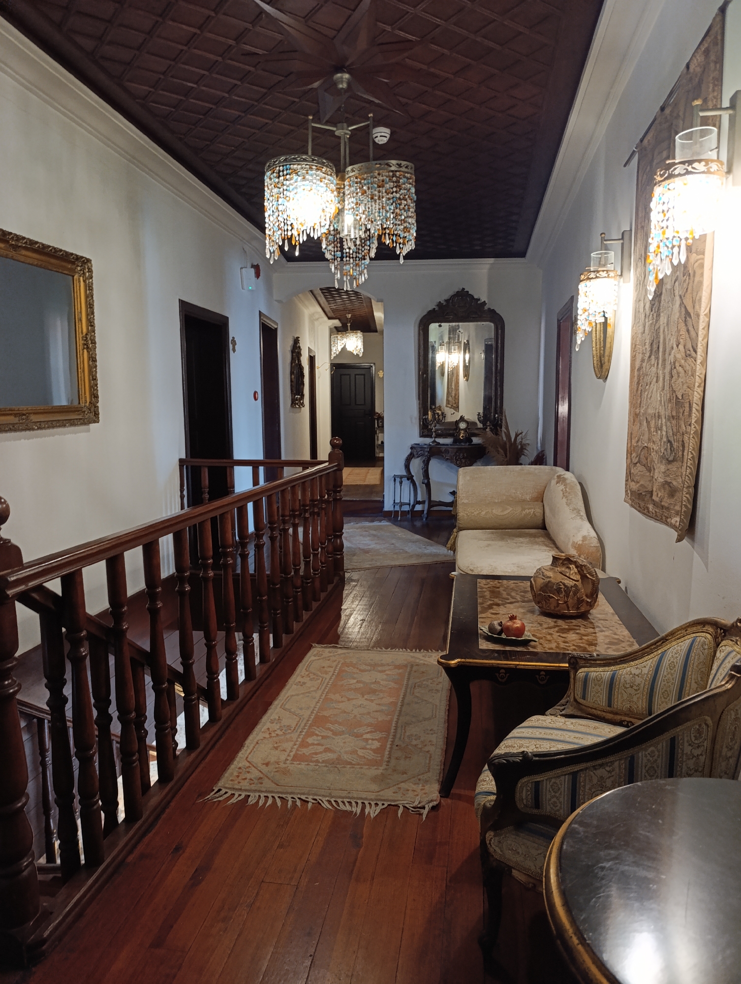The image depicts an ornate interior space, possibly a hotel or guesthouse, characterized by its historical and luxurious design. It is suggested that this location is in Antalya, Turkey.
The scene is a hallway, featuring dark wood flooring and a dark, coffered ceiling with white trim. A chandelier hangs from the center of the hallway, adorned with decorative crystals. Sconces with similar crystal details are mounted on the walls. The walls themselves are painted a bright white.
To the left, a wooden railing indicates a staircase leading downwards. There are a series of doorways leading to other rooms and spaces, including one at the end of the hallway.
On the right side of the hallway, various pieces of furniture are visible: a small couch, a table with decorative items, and an antique-style chair. A tapestry hangs on the wall near the couch. A mirror is placed at the end of the hallway.
The overall impression is one of opulence and old-world charm.