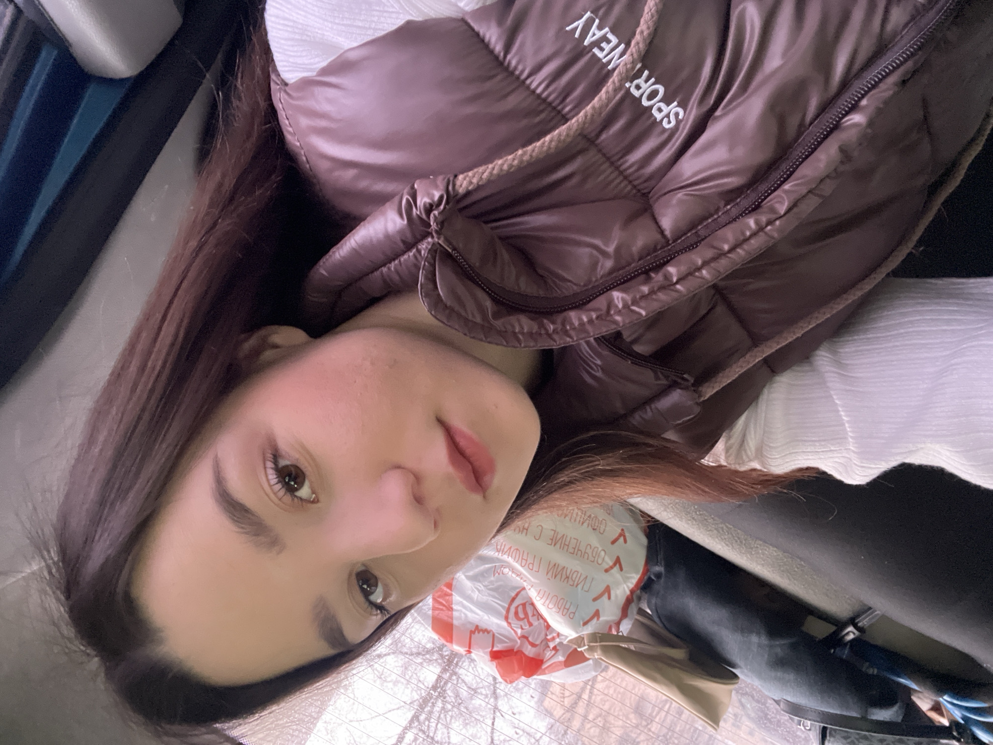 The image shows a young woman in a car, likely in Chaikovsky, Russia, as indicated by the Cyrillic text on the plastic bag. She is wearing a brown puffy vest with the word "SPORTWEAR" printed on the collar. Her dark hair is loose and styled. The background shows a window with bare trees outside. Part of the car's interior is visible. A shopping bag with red and white markings, partially obscured, rests near her.