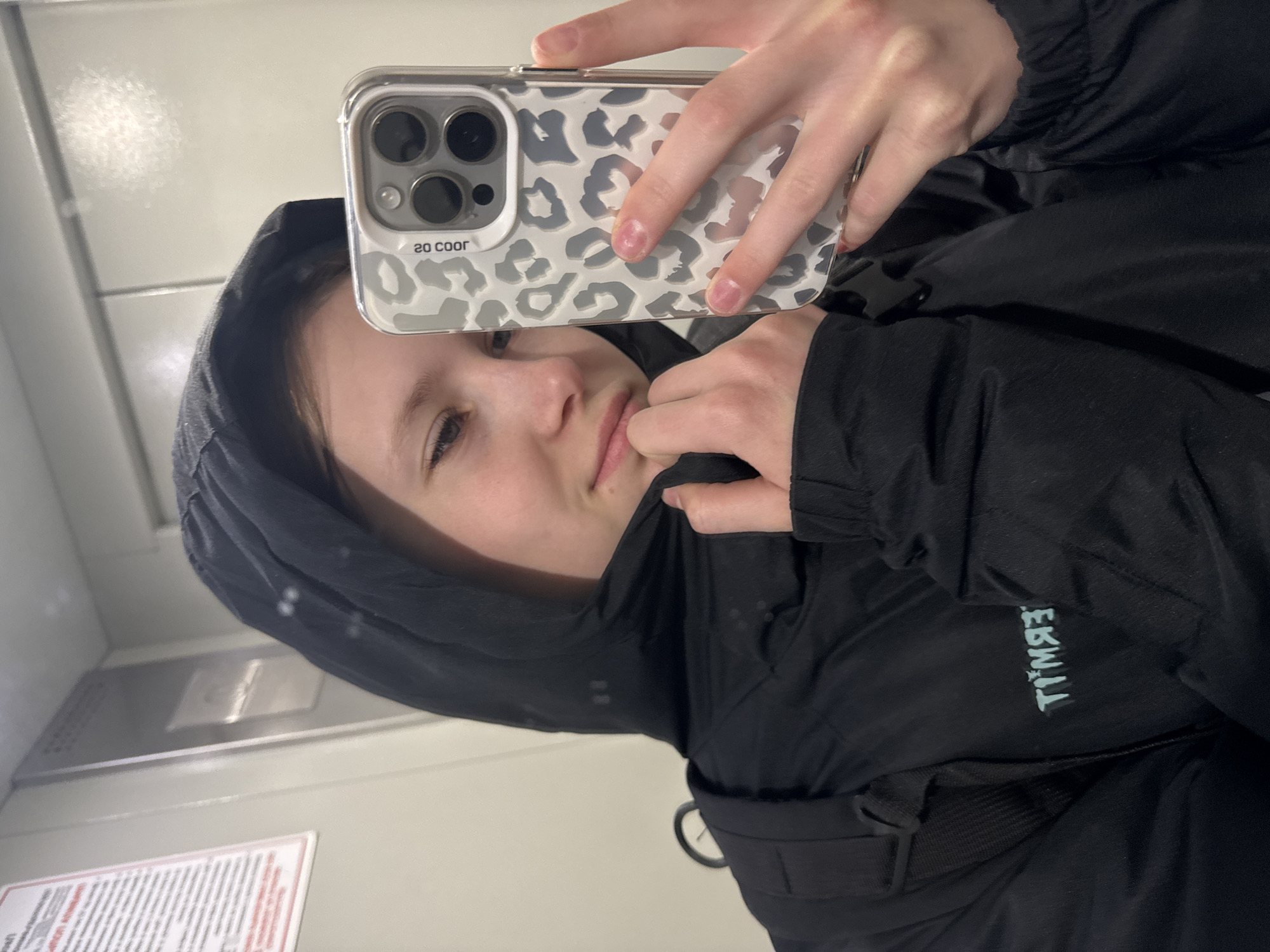 A person takes a selfie indoors, possibly on a train or bus due to the presence of a metal wall and informational sign in the background. The person is wearing a black hooded jacket. The person is holding an iPhone with a leopard print case to take the selfie. The brand 'Timewell' is visible upside-down on the jacket. The person's gender cannot be determined with certainty. The location in Chaikovsky, Russia, is speculative but cannot be confirmed from the image alone.