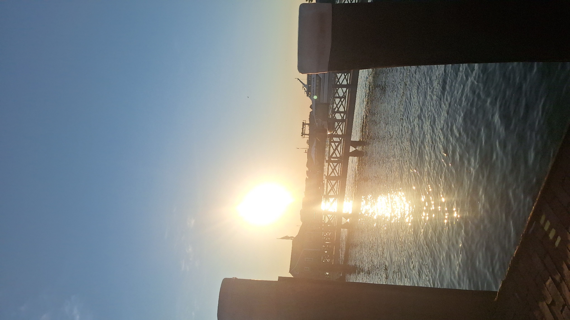 The image depicts a waterfront scene in Nantucket, USA, during what appears to be sunset or sunrise. In the foreground, two large, dark cylindrical pilings flank the view of a body of water. The water is rippled and reflects the bright sunlight, creating a shimmering effect. A wooden pier with a railing extends from the left side of the frame across the water. In the middle ground, several buildings with pitched roofs are visible along the shoreline, illuminated by the sun. Further in the background, various boats and structures, including what seems to be a ferry or larger vessel, are present. The sky is a clear, pale blue, with the sun positioned prominently in the upper left quadrant of the image, casting a bright glow and lens flare. The overall atmosphere is tranquil and scenic.