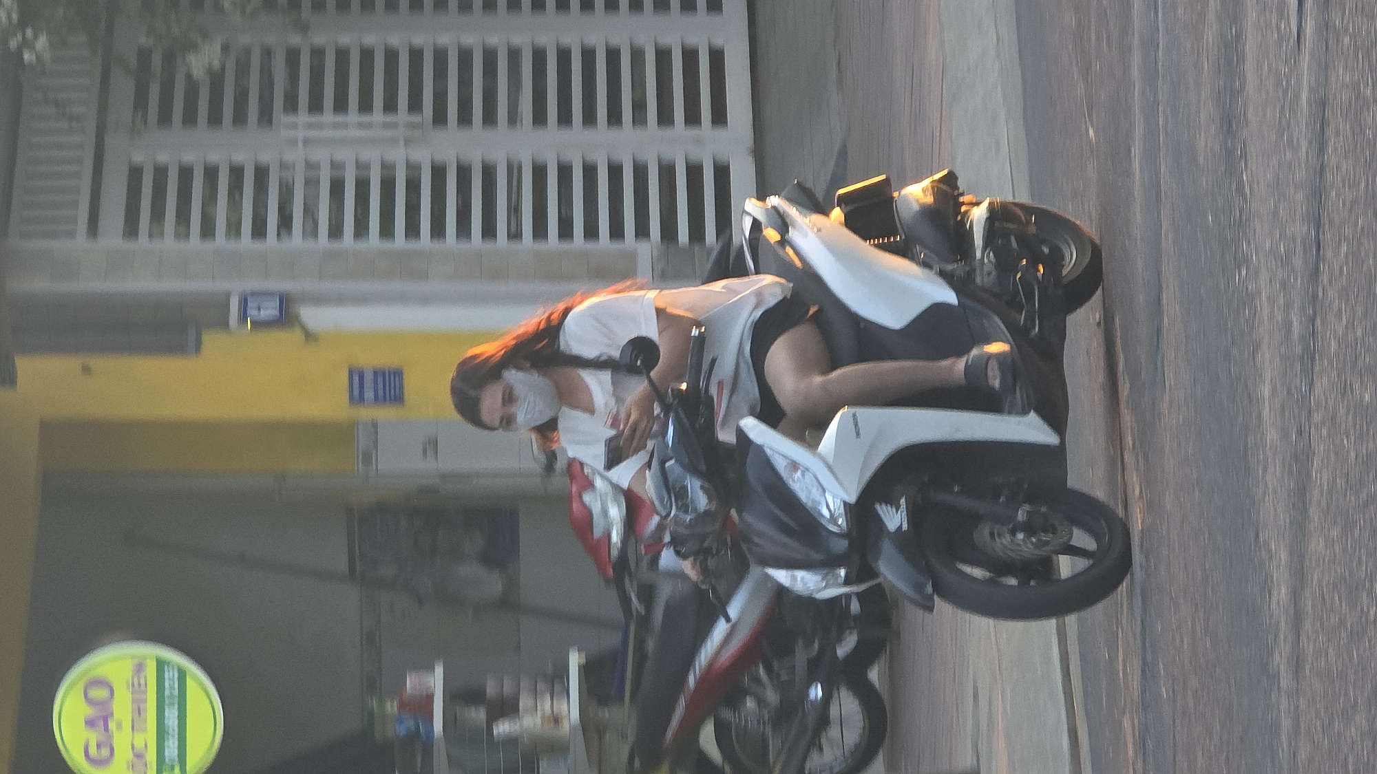 A single adult female is depicted riding a white and black Honda scooter on a paved road in Nha Trang, Vietnam. The woman has long, dark hair, wears a light-colored short-sleeved t-shirt, dark shorts, dark sandals, and a light gray face mask. She is actively operating a white smartphone with both hands while maneuvering the scooter. The scooter's front fender displays a Honda logo. In the background, to the right, is a yellow building with a white slatted double gate, and a blue street number sign displaying "17" is affixed to the yellow wall. To the left, a building storefront is partially visible with shelves containing items and a circular green sign for "GAO DUC THIEN" (rice brand) with a phone number. Several parked motorcycles, including a red and black model, are visible behind the rider. The scene is illuminated by natural light.