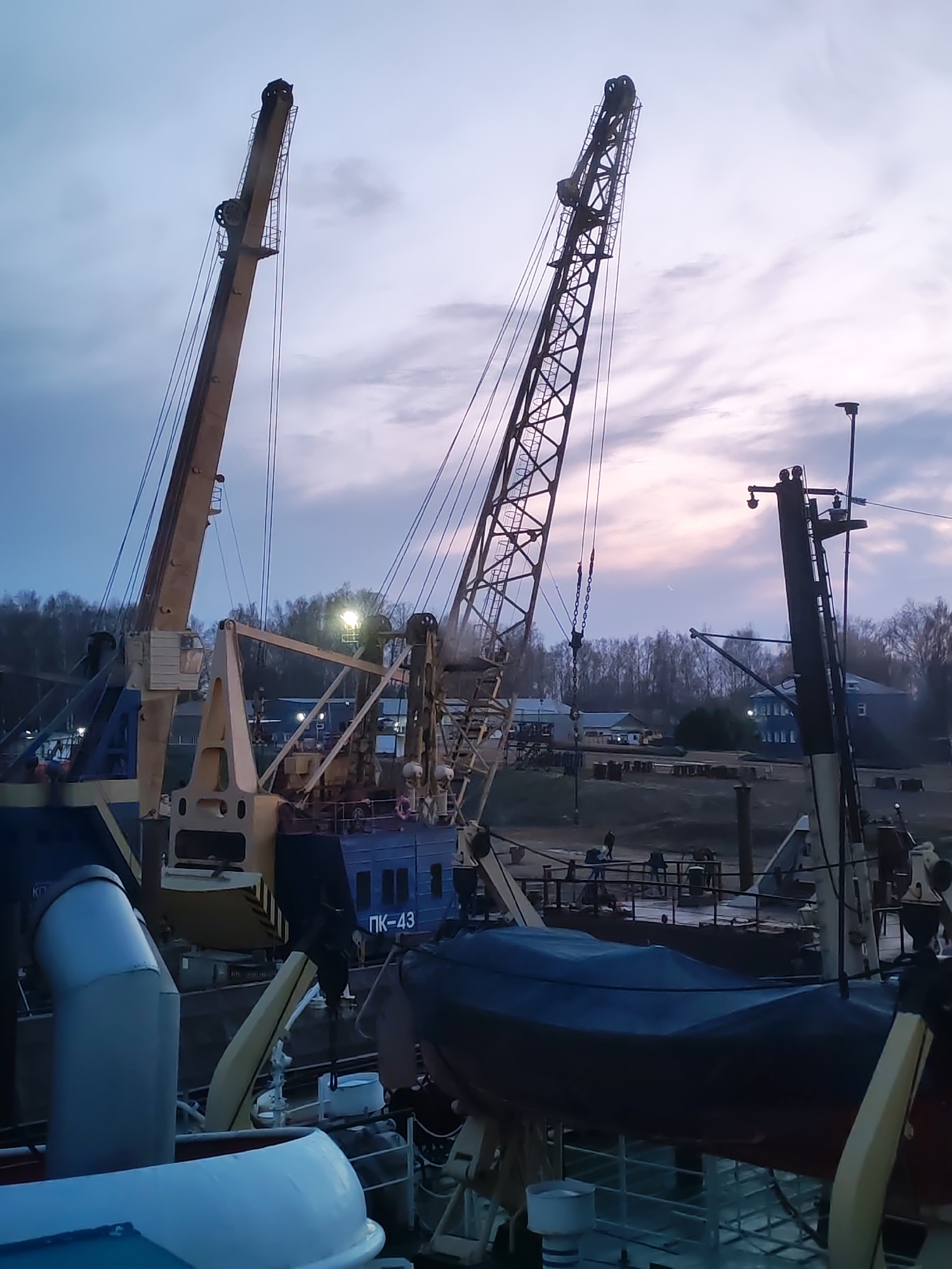 The image captures a view from what appears to be the deck of a ship or barge, overlooking a waterfront in Rybinsk, Russia. The scene includes two prominent cranes mounted on vessels. The crane on the left is painted in light colors, and the one on the right has a lattice structure, characteristic of a crane boom. Another, smaller crane is also visible on the right of the image, close to a mast with attached equipment.
On the deck of the vessel from which the photograph is taken, a covered lifeboat is visible. In the background, on the other side of the water, there are buildings, trees, and dockside equipment, suggesting an industrial or transport hub. Several figures can be seen walking along the dock. The sky suggests it is either dusk or dawn with pastel-colored clouds.
