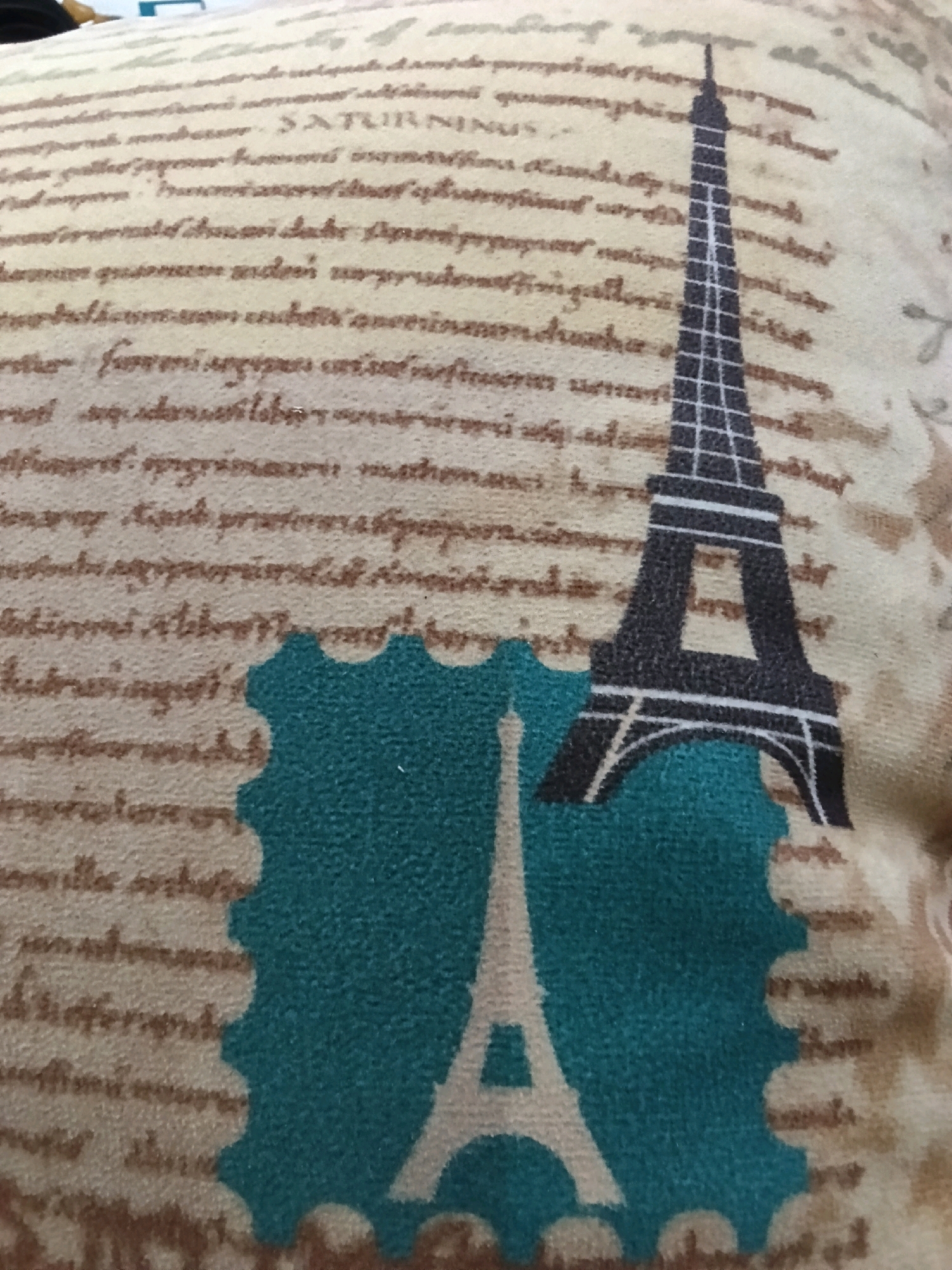 The image displays a close-up of a fabric surface, likely a pillow or tapestry, featuring a design reminiscent of old documents and Parisian landmarks. The background is a light beige with a textured appearance, overlaid with faint, cursive-style text in a darker brown hue. Prominently featured in the center is a silhouette of the Eiffel Tower, rendered in black and white. The tower is superimposed on a teal-colored, scalloped rectangular shape that resembles a postage stamp. The bottom portion of the image shows a smaller, lighter-colored silhouette of what appears to be another Eiffel Tower against the teal stamp. The overall aesthetic evokes a vintage travel theme. The location of Bennane, Tunisia is not depicted or suggested by any elements within the image.