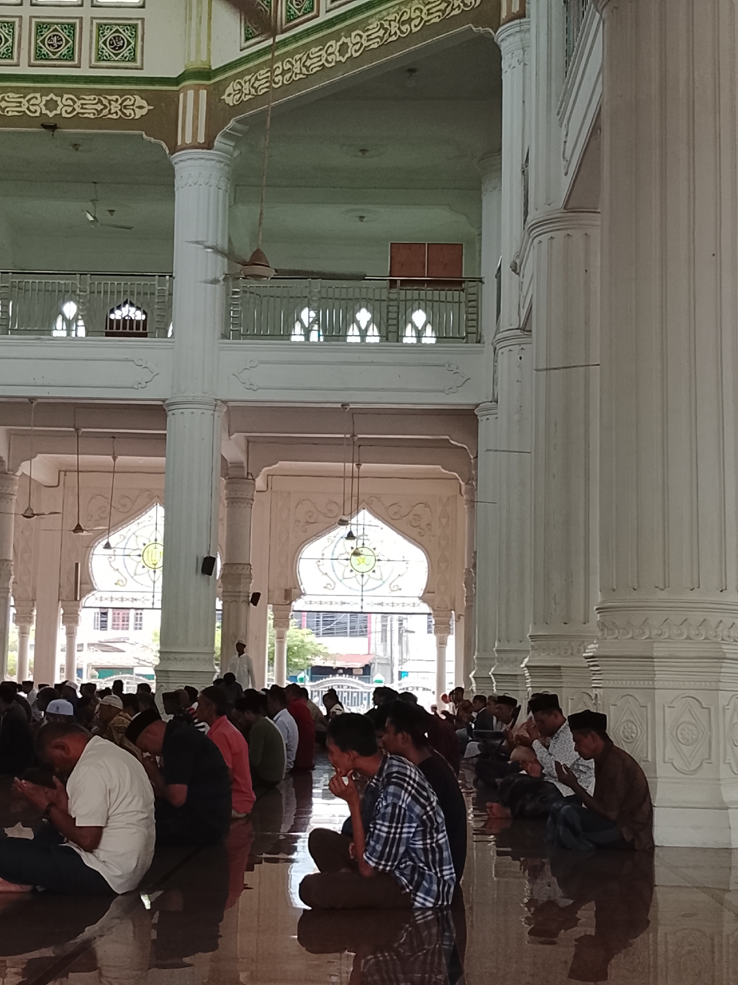 The image shows a group of men sitting on the floor of a mosque, in prayer. The mosque is large, with tall white pillars and ornate arches. Sunlight streams in through stained-glass windows, illuminating the polished floor. The men are dressed in traditional Islamic attire, with some wearing prayer caps. The scene is peaceful and contemplative. The mosque is likely located in the city of Lhokseumawe, Indonesia, given the architectural style and the presence of Muslim worshippers.