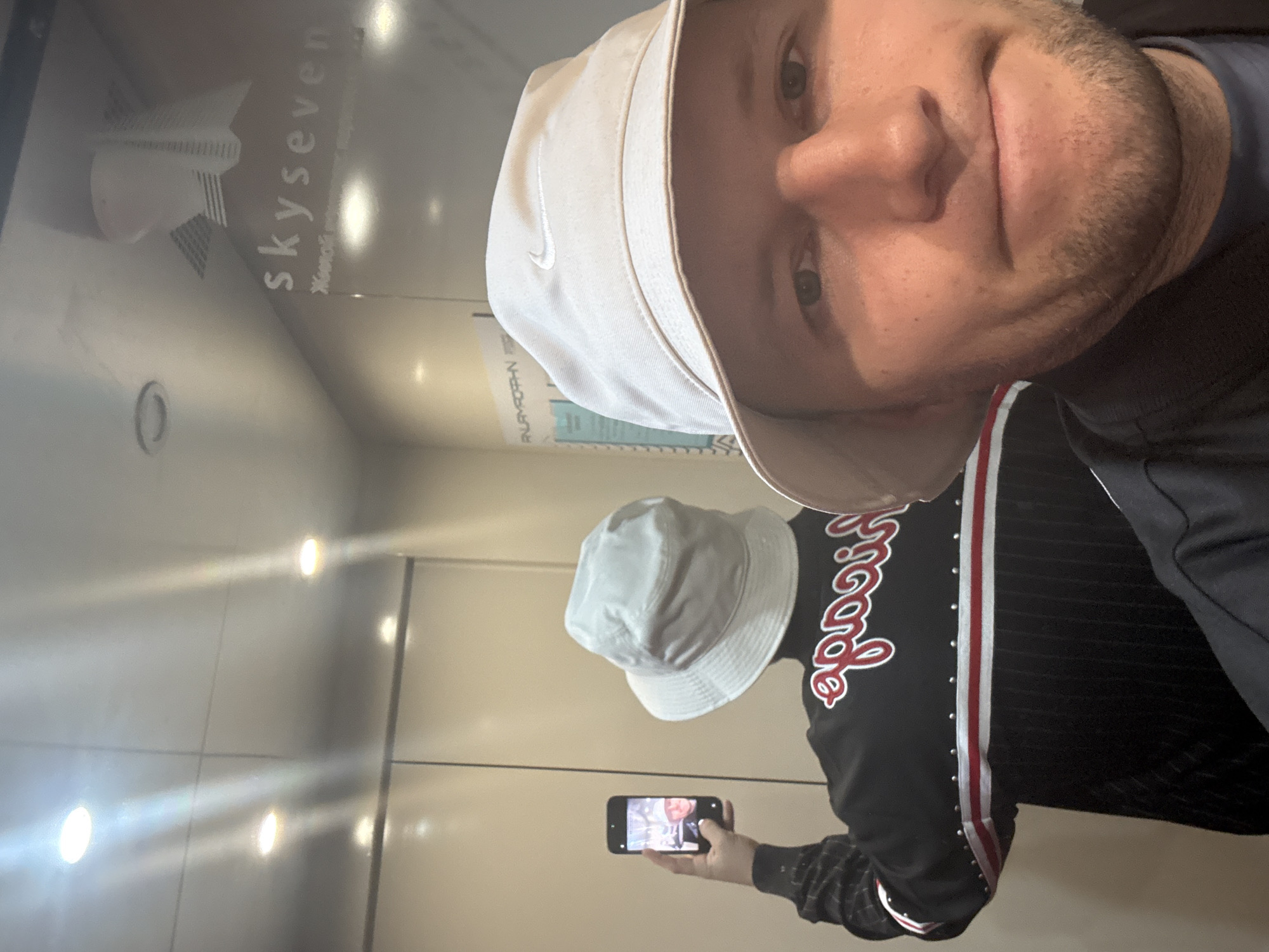 A man is taking a selfie in what appears to be an elevator. He is wearing a light-colored bucket hat with a Nike logo and a black jacket with a red and white stripe down the side. In the reflection of the elevator door, he is taking a picture with his smartphone. The sign on the wall reads "skyseven" and "Жилой комплекс в сердце". The scene is likely within a residential complex in Krasnoyarsk, Russia. The elevator walls are a light color.