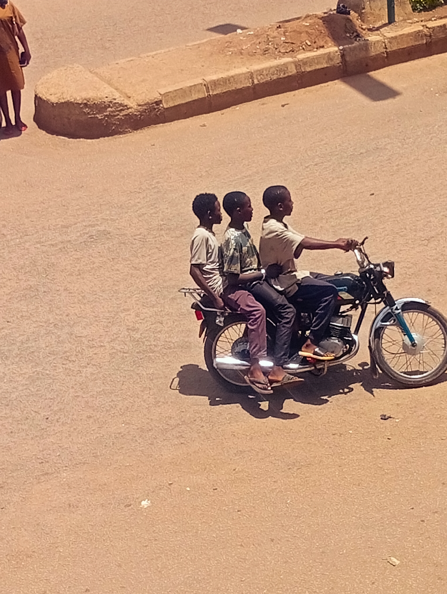 Three young Black males are on a motorcycle moving right on a brown asphalt road. The driver wears a light tan short-sleeved shirt and dark pants, with brown sandals. The second male wears a green patterned t-shirt, dark pants, and brown sandals. The third male wears a light tan t-shirt and red pants, with brown sandals. The motorcycle is dark teal with silver accents. There is a woman to the left of the frame walking away from the camera wearing a tan dress. There is a curb on the right side of the frame with greenery behind it. The location is a city in Unknown, Unknown.