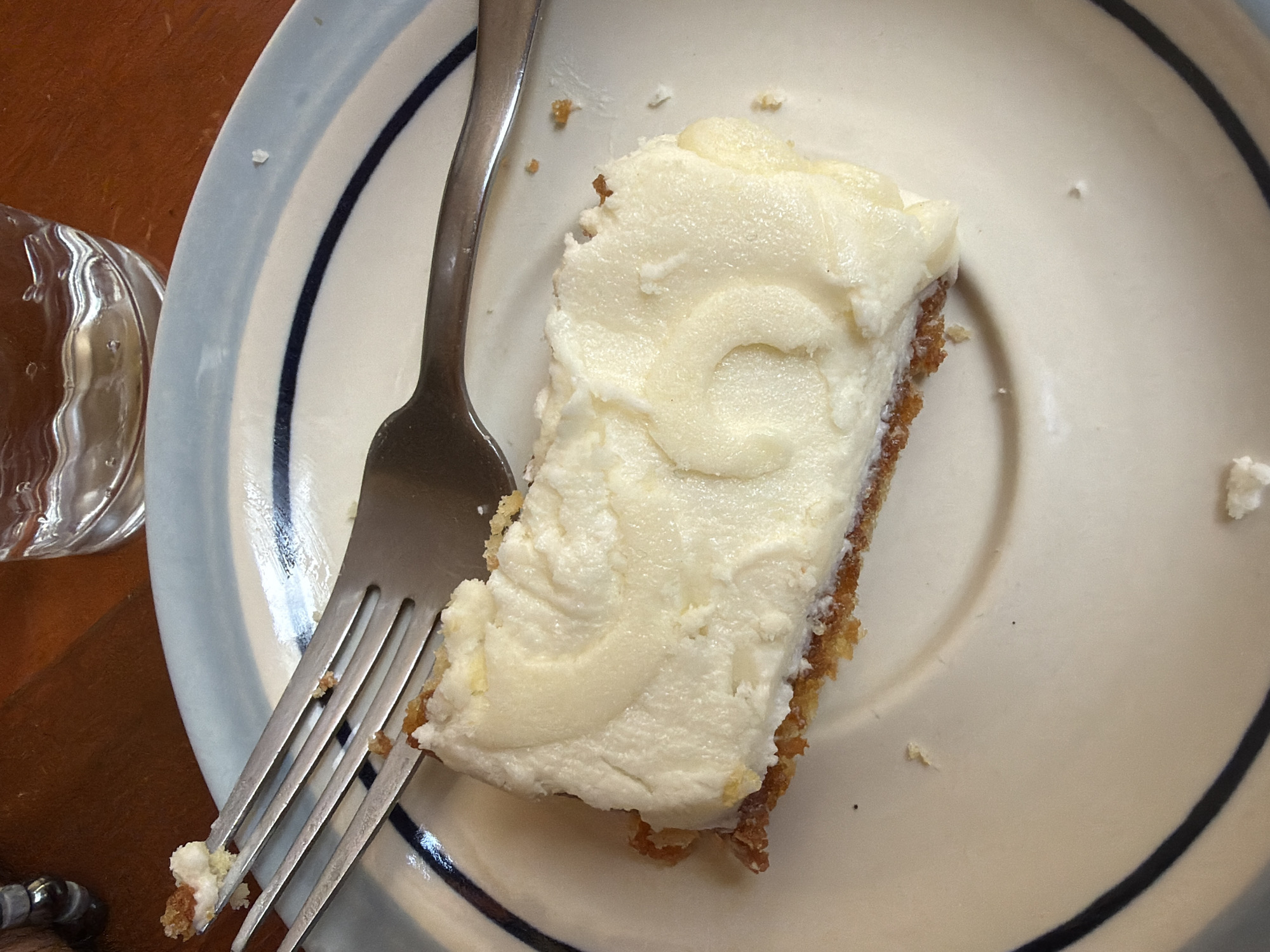 A close-up, overhead shot depicts a partially eaten slice of cake on a white plate with blue rings. A silver fork rests on the plate, with cake crumbs caught in its tines. The cake has a crumbly base and a thick layer of white frosting. A glass of water is visible in the lower left corner, next to a wooden table. The specific location is unknown.