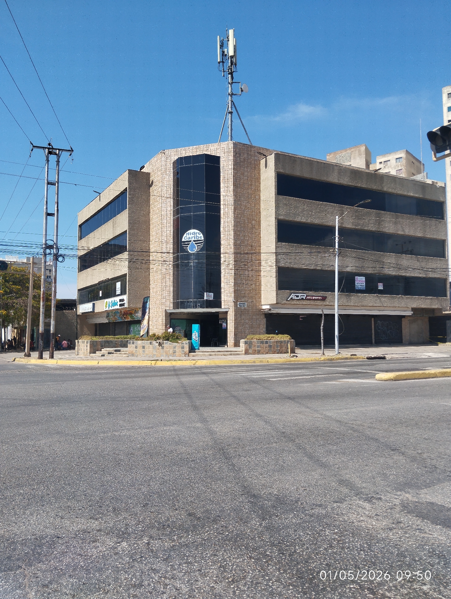 The image shows a street scene in Porlamar, Venezuela, on January 5, 2026, at 09:50.
Dominating the frame is a four-story commercial building. It features a tan brick facade with horizontal bands of dark-tinted windows. The central section of the building is a recessed glass facade, with a "Hidro Caribe" logo visible on the glass. A cellular tower is mounted on the roof.
At street level, there are several businesses. "ADR Auto Partes" is visible. Several storefronts appear to be closed. "Se Venden" signs are posted.
In the background, there are other buildings, utility poles, and trees. The sky is clear blue.
The foreground is dominated by a wide asphalt street. Sidewalks with low retaining walls are visible along the building's perimeter. A few people are seated on the sidewalk to the left. A traffic light is visible in the right background.