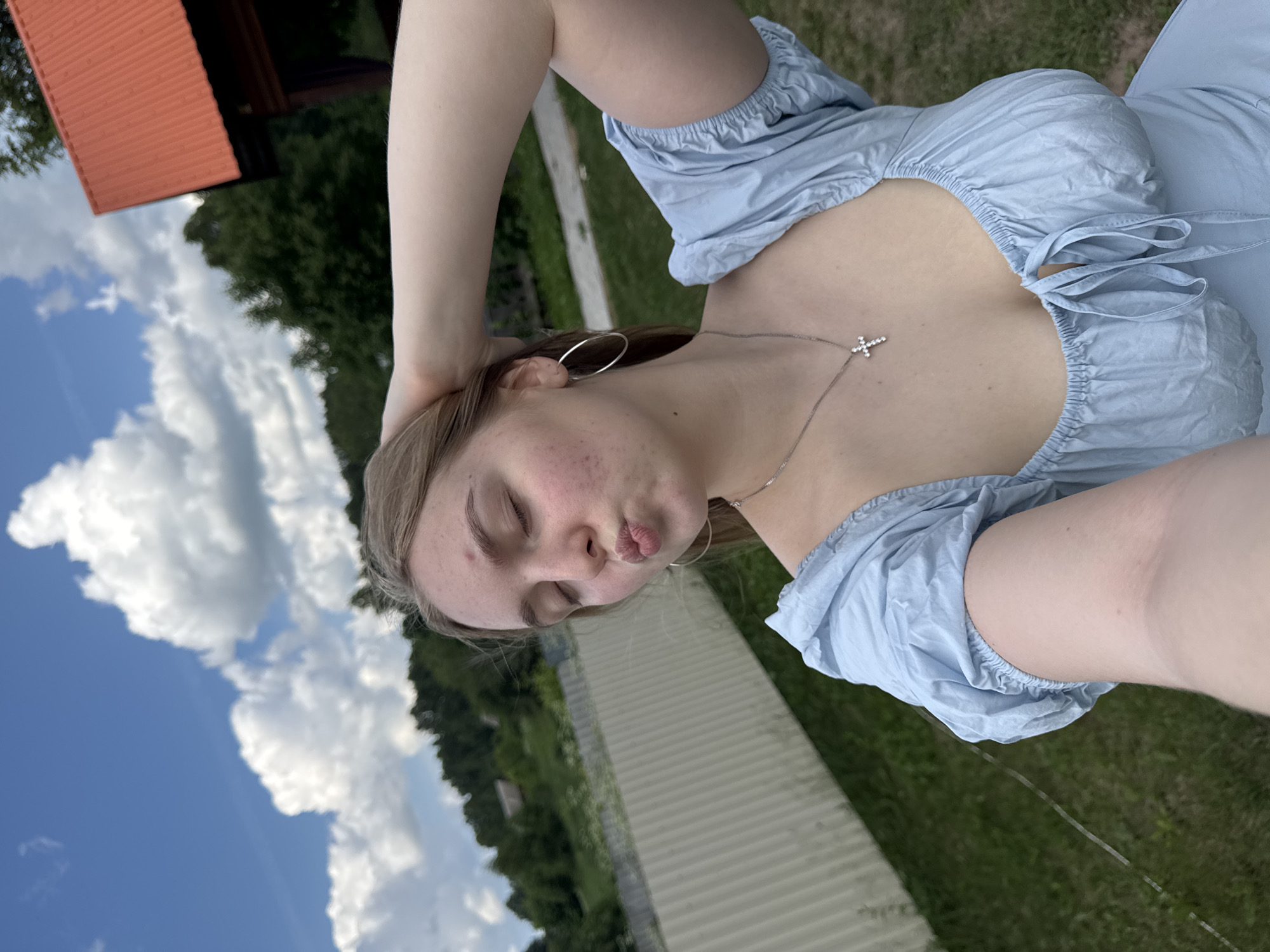 In a low-angle, outdoor selfie, a young woman with blonde hair poses with her eyes closed and lips puckered, her right hand behind her head. She wears a light blue dress with puff sleeves and a tie front, accessorized with hoop earrings and a cross necklace. The background suggests a rural setting in Russia, with a white fence, green grass, trees, and a blue sky dotted with white clouds. To the upper right, a structure with an orange roof is partially visible. The image is taken in the city of Unknown, Russia.