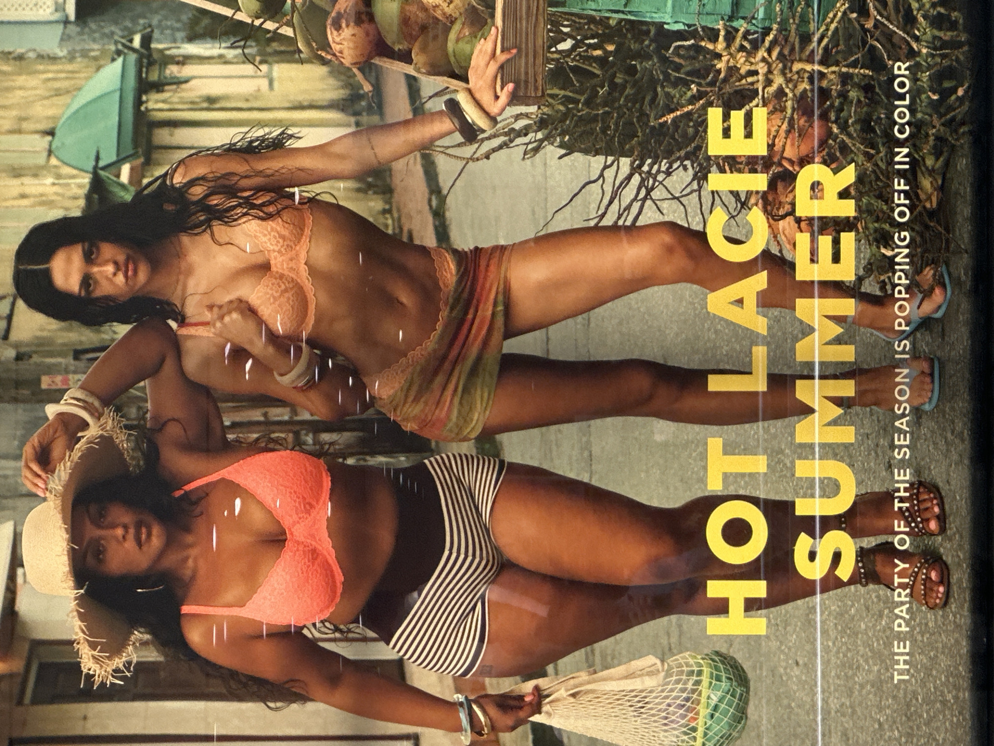 This image appears to be an advertisement featuring two women posing in lingerie or swimwear, positioned against an urban backdrop. The woman at the bottom of the image is wearing a straw hat, a bright pink lace bra, and black and white striped shorts. She carries a netted bag. The woman at the top of the image is wearing a peach-colored lace bra and a sarong-like garment. She is near a display of coconuts. The text "HOT LACIE SUMMER" is superimposed on the image. The text "THE PARTY OF THE SEASON IS POPPING OFF IN COLOR" is included in smaller print. Due to the limited visual information, it is not possible to confirm the location as Burnaby, Canada.