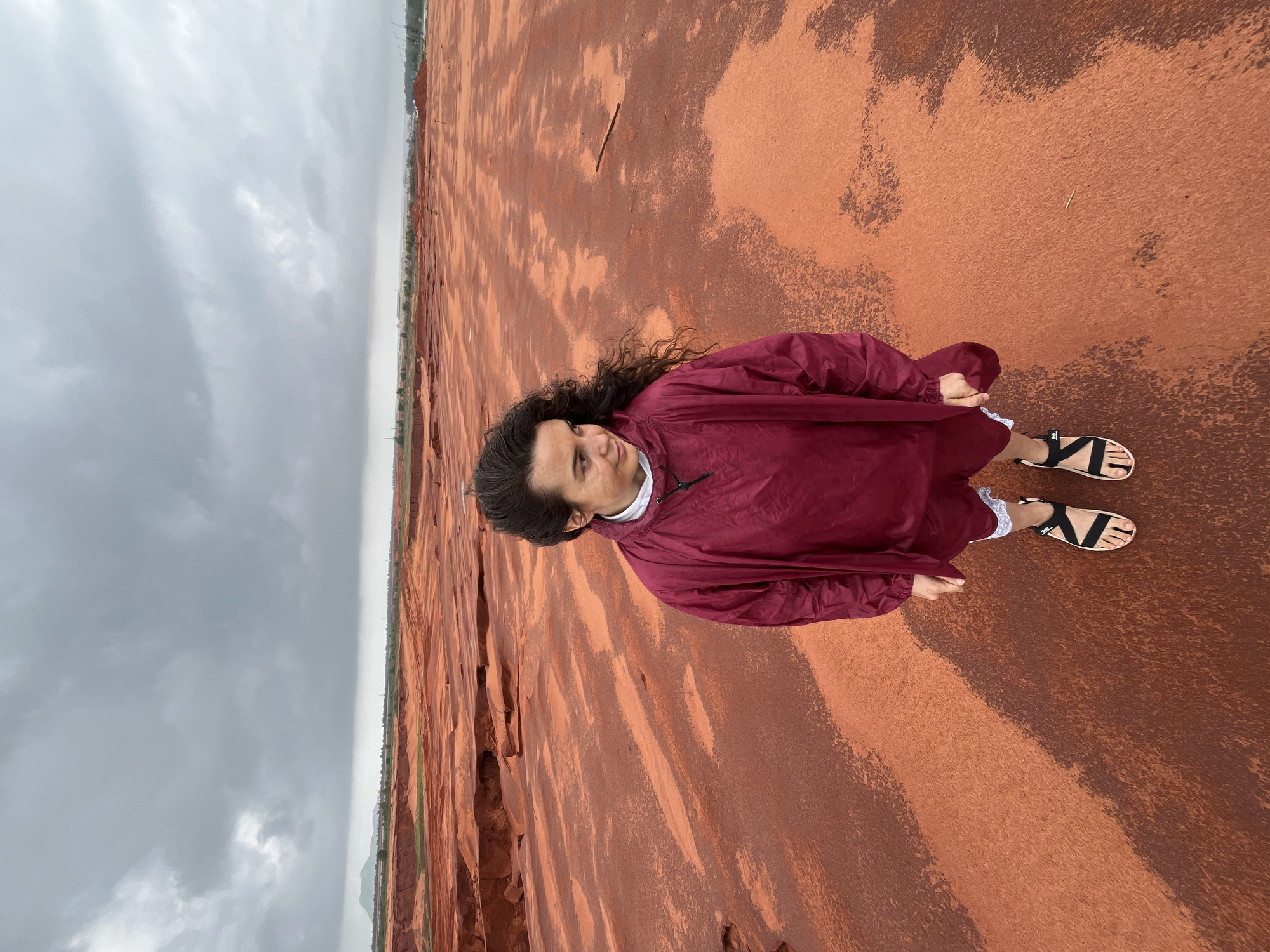 A woman stands in the center of a vast expanse of reddish-orange earth under a cloudy sky. She is looking to her right, her long, dark, curly hair framing her face. She is wearing a maroon windbreaker jacket over a white top and maroon shorts with white trim. On her feet are black and beige sandals. The ground appears uneven, with some areas lighter and some darker, suggesting it is damp in places. In the background, the landscape is flat with some distant trees and hills, and a faint indication of a horizon line where the land meets the sky. The sky is overcast with heavy grey clouds, hinting at impending rain or a recent downpour. The overall impression is one of a desolate, open landscape. The location is identified as Mui Ne, Vietnam, a region known for its sand dunes.