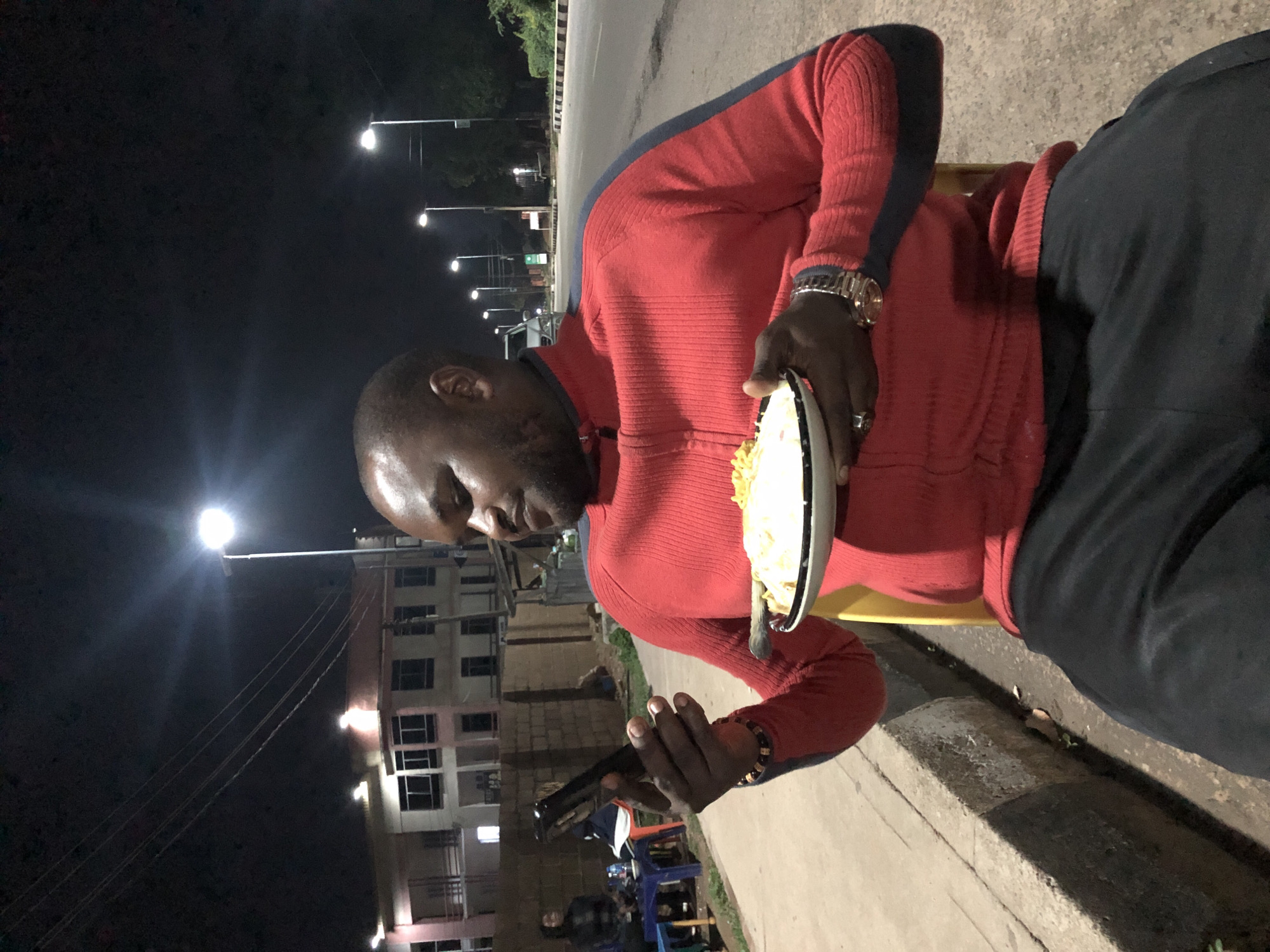 In a nighttime scene on a city street in Jos, Nigeria, a man sits in a yellow plastic chair holding a plate of food in his right hand and a smartphone in his left hand. He is wearing a red zippered sweater with dark blue accents on the sleeves and collar, and black pants. A wristwatch with a rose gold-colored band is visible on his left wrist. He appears to be looking at his phone as he holds the plate. The background shows a street with multiple streetlights, buildings, and a sidewalk. The lighting suggests it is after dark, with artificial light sources illuminating the area. There are some other people and objects visible in the background, indicating a public space.