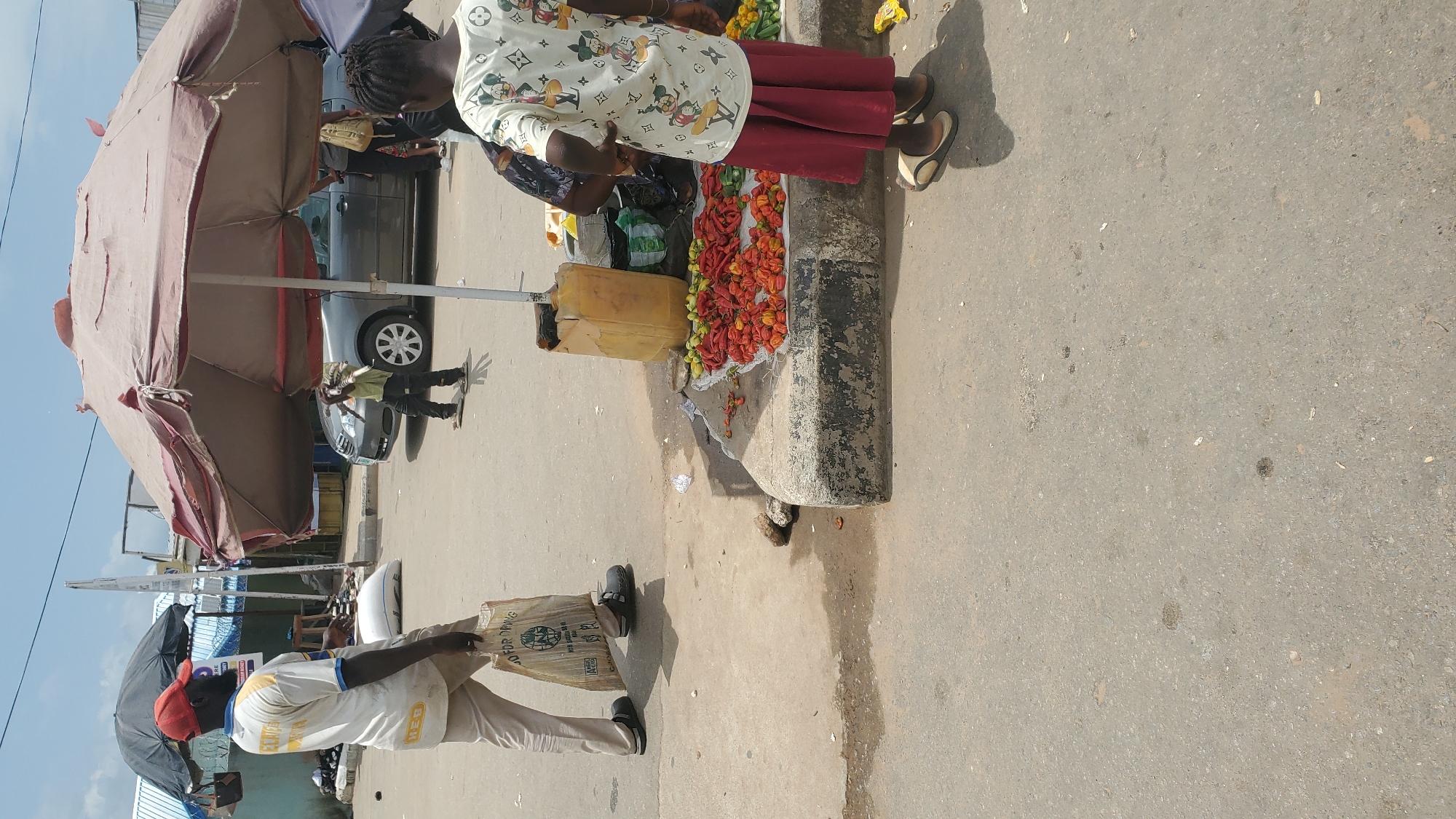 The image depicts an outdoor market scene in Jalingo, Nigeria. In the foreground, a person wearing a light-colored t-shirt with cartoon characters and a red skirt is standing. To their right, a large umbrella shades a stall where red and yellow peppers are displayed on the ground, next to a yellow plastic container. A man in a white t-shirt and khaki pants is walking away from the viewer, carrying a woven bag. In the background, a gray car is parked, and other market stalls and structures are visible. The ground is paved with asphalt and concrete. The sky is partly cloudy.