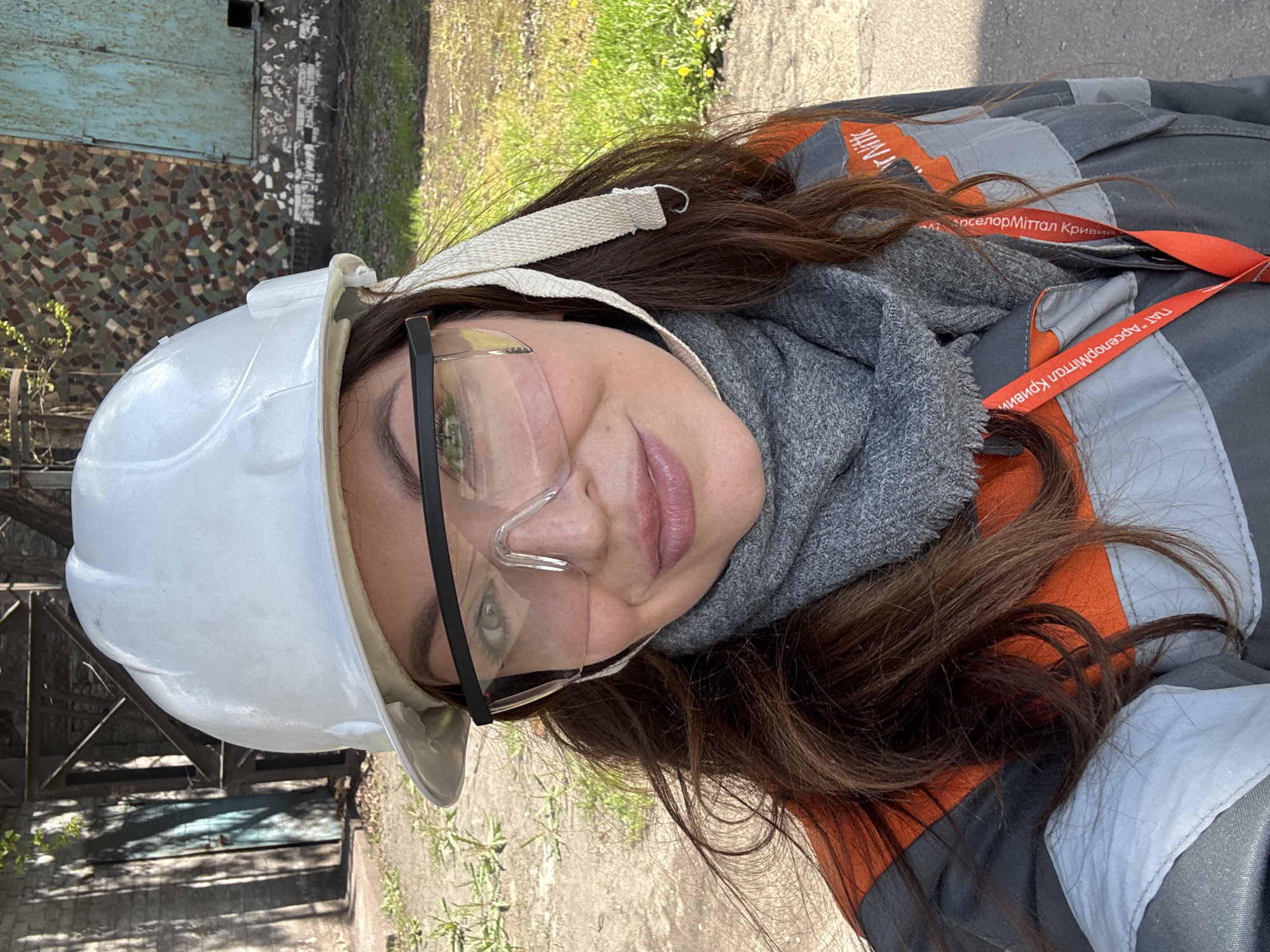 A woman is posing for a selfie in an industrial area. She is wearing a white hard hat, safety glasses, and a grey scarf. Her work clothes are grey and orange, and a red lanyard with the name "АрселорМиттал Кривий Ріг" (ArcelorMittal Kryvyi Rih) is visible. The background includes a building with a mosaic pattern and a concrete area with grass. The photo was likely taken in Kryvyi Rih, Ukraine, given the text on the lanyard.