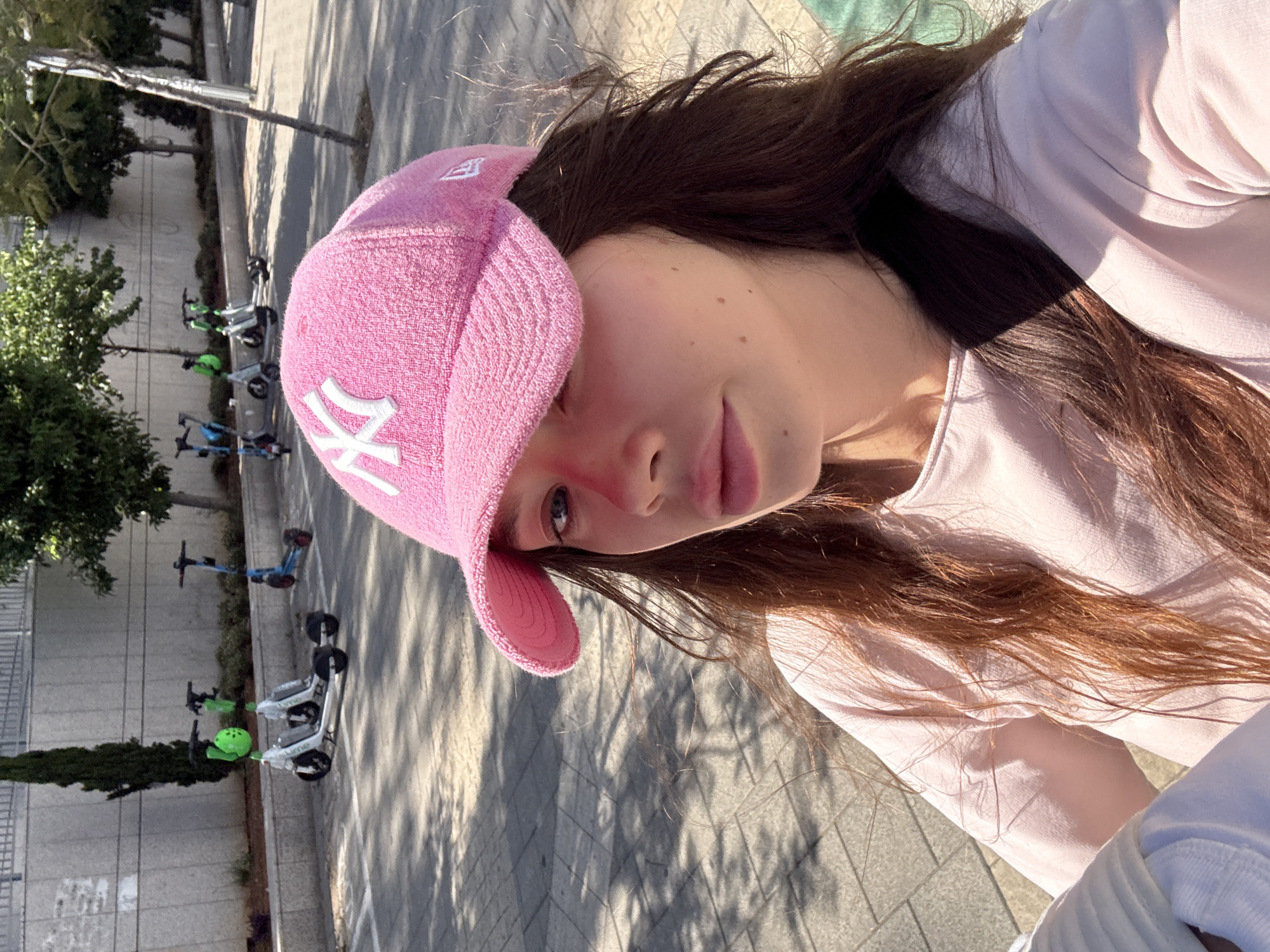 A young woman wearing a pink New York Yankees baseball cap and a light pink shirt stands outside on a sunny day. A line of rentable electric scooters is parked against a building in the background. It is not possible to specifically verify the location in Tel-Aviv, Israel.