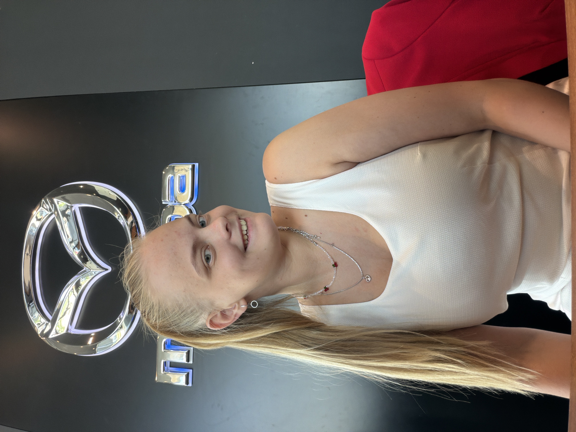 A young woman is standing in front of a Mazda logo. She is wearing a white top and a necklace. She has long blonde hair. There is a red object behind her. The image may have been taken in Izhevsk, Russia, though there is no visible evidence to confirm this.