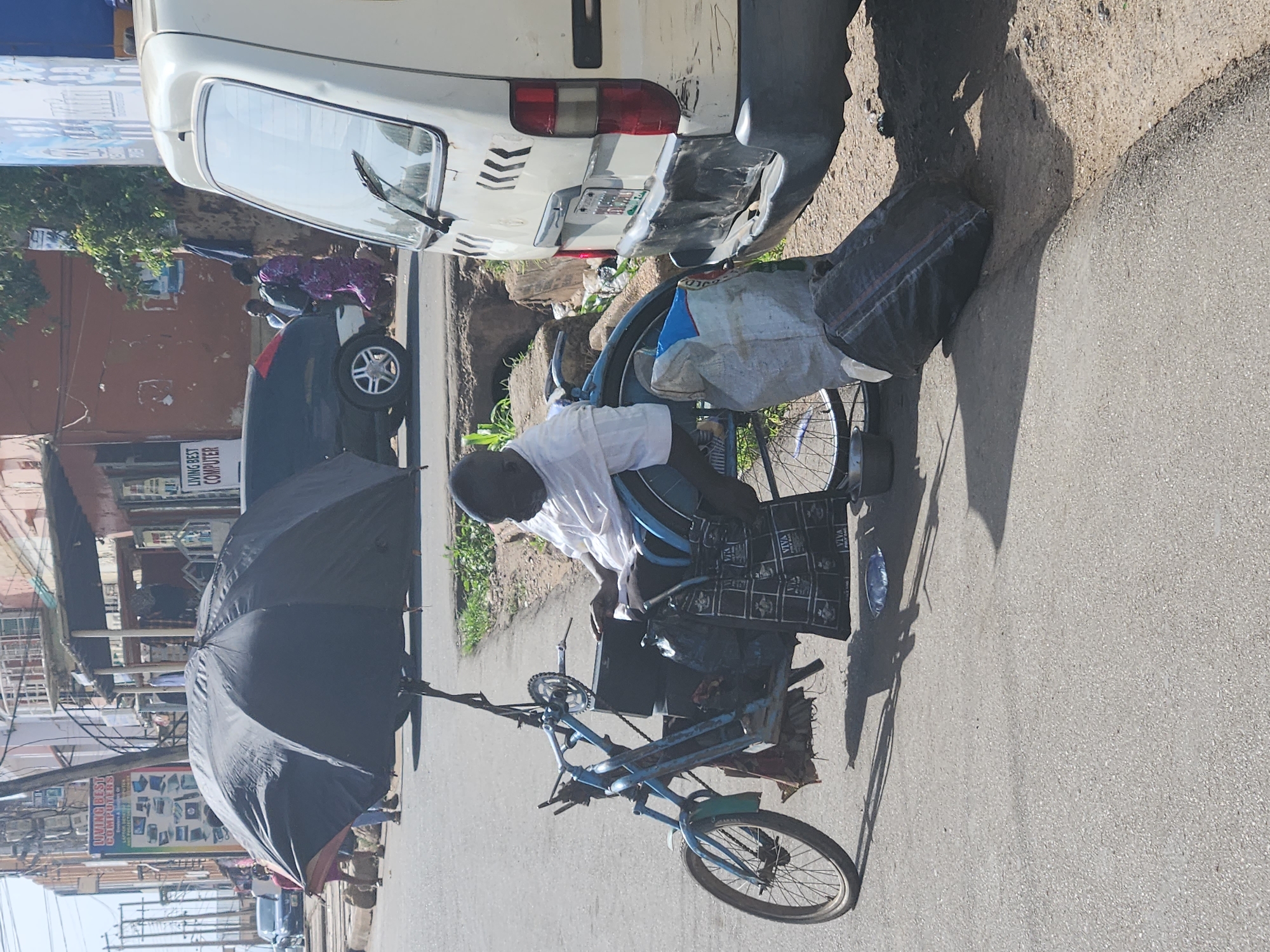 The image depicts a man in a white shirt riding a blue tricycle-like vehicle with an umbrella attached, along a street in an urban environment. The street is paved and adjacent to a sidewalk with vegetation. A white van is parked nearby, partially obstructing the view. Buildings, signage, and parked cars are visible in the background. The image is taken during daylight with clear weather.