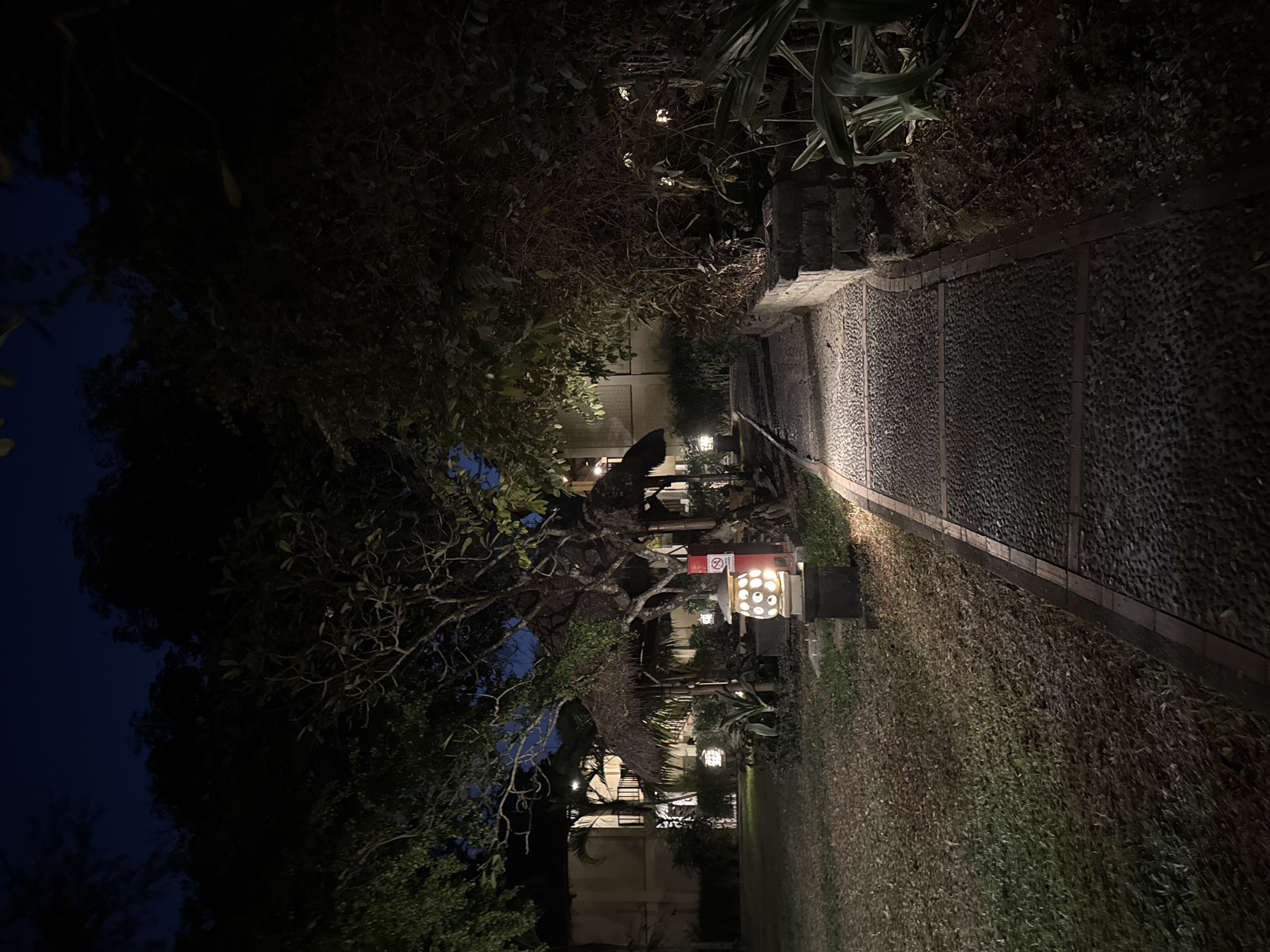 The image depicts a dimly lit pathway at night, likely in a resort or hotel in Denpasar, Indonesia. The pathway is paved with dark stones and bordered by grass on the left and a stone wall with lush vegetation on the right. Several decorative lanterns are strategically placed along the path, casting a warm glow. In the background, a building with balconies and thatched-roof structures suggests a tropical setting. The overall atmosphere is tranquil and serene, evoking a sense of peace and relaxation.
