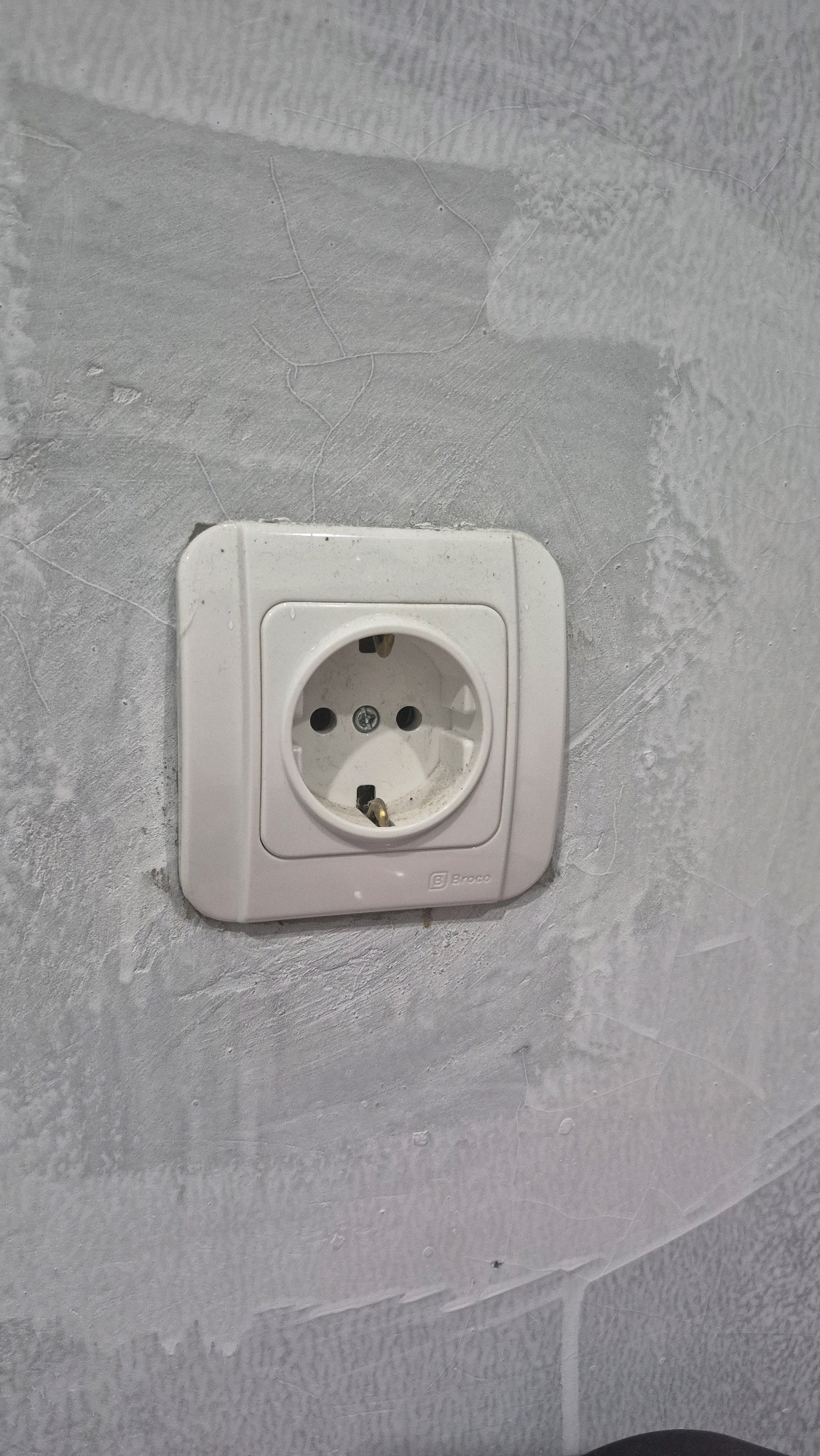 A white electrical outlet is mounted on a grey, textured wall. The outlet is a standard European type with two round holes and a grounding pin. The wall appears to be unfinished or under renovation, with visible patches of different textures and some hairline cracks. The outlet has a logo that reads "Broco". The image is taken from a medium-close-up perspective, focusing on the outlet and the surrounding wall. There are no people or other significant objects in the frame. The lighting is even, suggesting an indoor setting. The location is specified as Gambiranom, Indonesia.