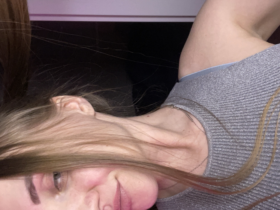 A woman's tilted face is visible with a silver, ribbed top. Her light brown hair partially covers her face. Her left eye is open and looking downwards, while her lips are closed. Her complexion is fair, and a delicate silver necklace graces her neck. The background appears dark with a faint pink glow near the top left corner.