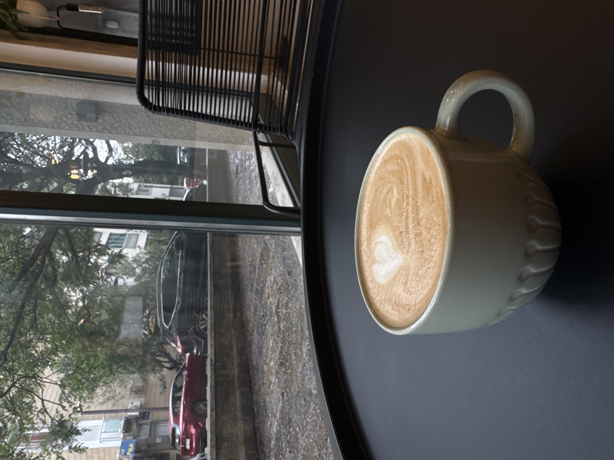 A top-down, medium shot depicts a white ceramic mug of cappuccino with latte art on a dark gray table next to a window. Outside the window is a view of an urban street with buildings, trees, and parked cars; possibly Budva, Montenegro. There is a black metal chair partially visible in the upper-left corner of the frame. No people are visible in the scene.
