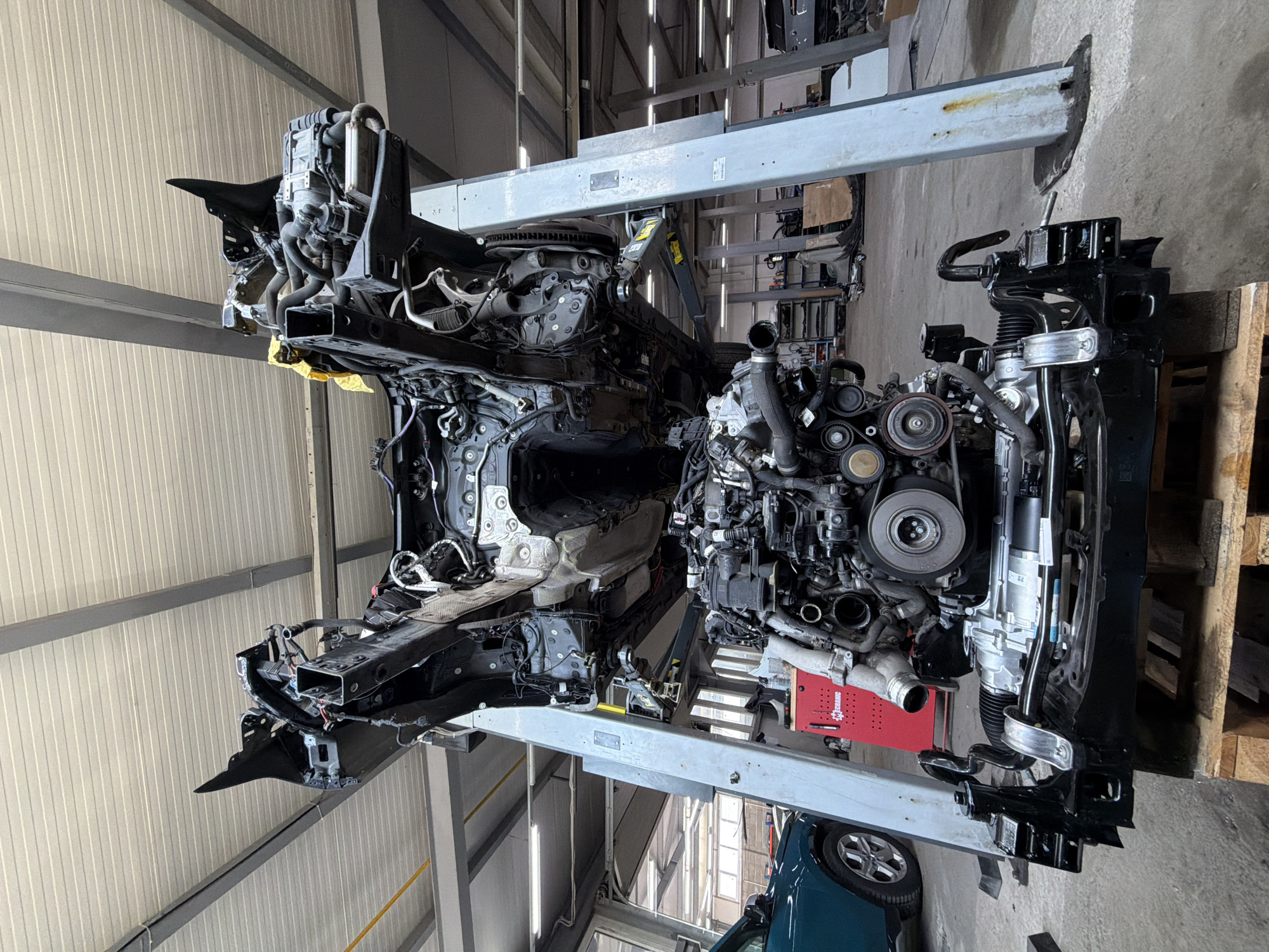 The image shows the disassembled front end of a vehicle suspended on a lift inside what appears to be an automotive repair shop. The engine and other components are detached from the chassis and are also suspended. The scene seems to be an indoor location with an industrial interior: metal walls, structural beams, and other equipment are visible in the background. The location is unspecified, and there's nothing in the image that would explicitly indicate the location as Burgas, Bulgaria.