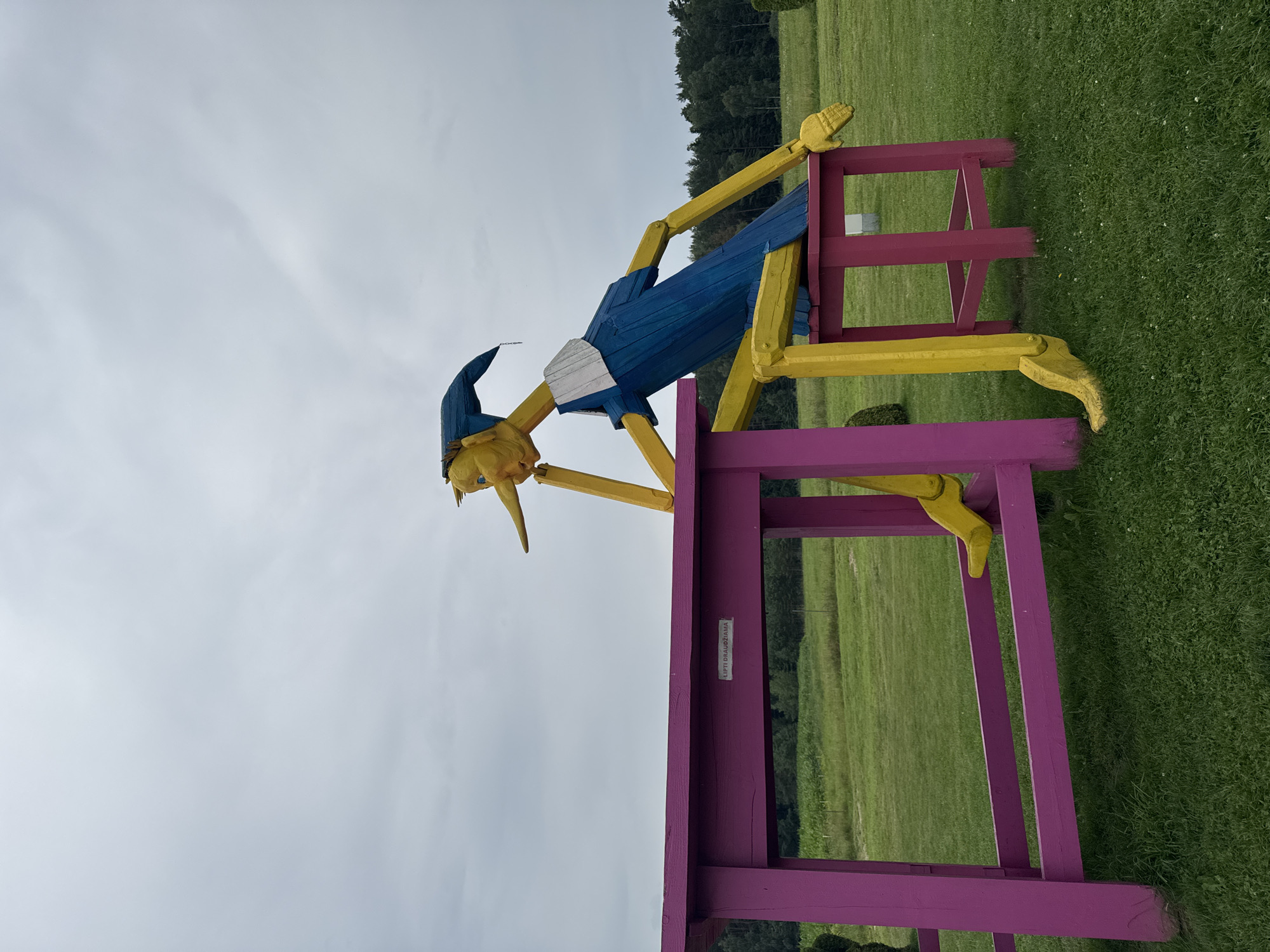 In Skapiškiai, Lithuania, a large wooden sculpture of a whimsical figure, resembling Pinocchio, sits at a pink table and stool. The figure has a yellow body with blue clothing and a blue hat. Its long nose and facial features are prominent. The figure is posed as if deep in thought, with one arm resting on the table. The table and stool are bright pink and are set outdoors in a grassy field. In the background, there are trees and a cloudy sky. A sign on the table reads "LIPTI DRAUDŽIAMA," which translates to "Climbing is forbidden."