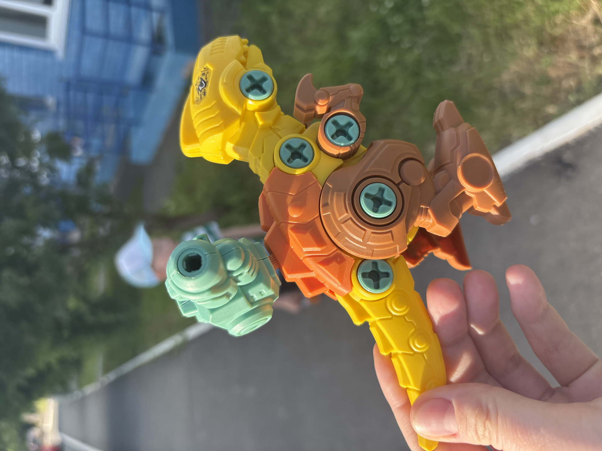 An outdoor, sunlit, eye-level, medium shot features a person's hand holding a disassembled, multi-colored toy dinosaur. The dinosaur is composed of yellow, orange, and brown plastic parts that are held together by teal-colored screws. A green, robotic-looking head is attached to the top of the dinosaur's body. In the blurred background, a person wearing a light blue cap and a striped shirt is visible, standing on a paved path next to a grassy area. A blue building with white trim and windows is also present in the background. The scene appears to be located in Izhevsk, Russia.