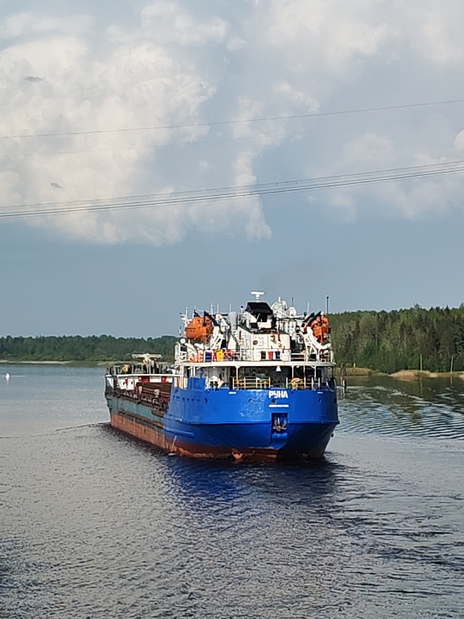 A blue and red cargo ship labeled "PYHA" is navigating a waterway, likely a river or canal, near Gari, Russia. The ship is moving away from the viewer. Its deck is white with various structures and orange life rafts. Dense green forest lines the shores. The sky is partly cloudy with visible power lines extending horizontally across the upper part of the frame. The water is dark with visible ripples.