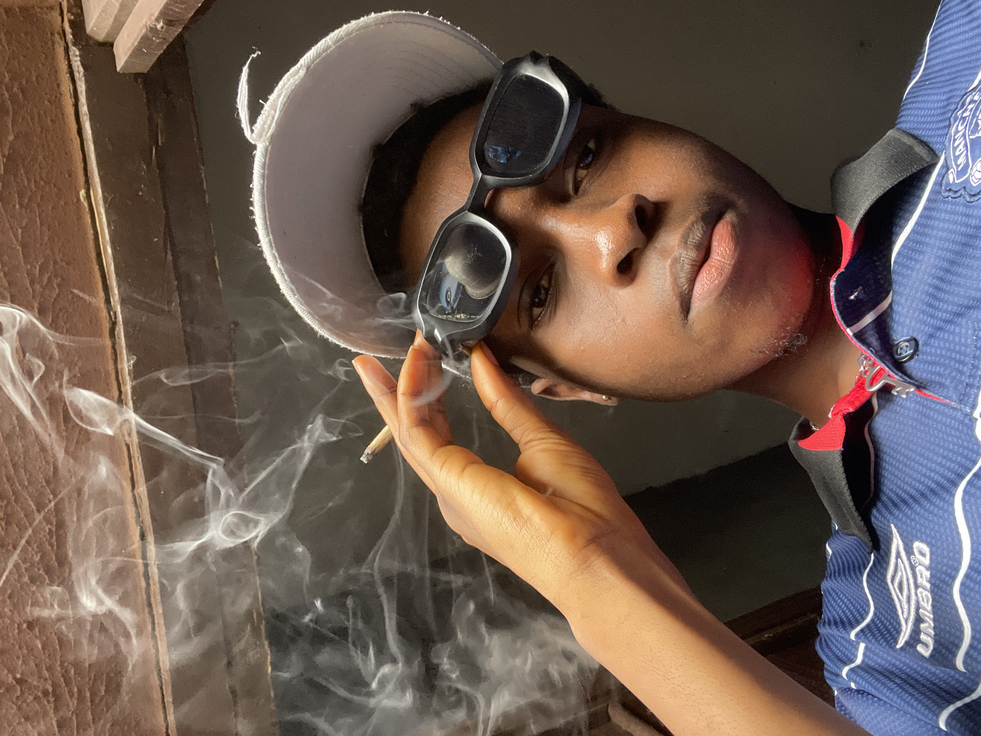 The image shows a young man with dark skin wearing a white baseball cap and rectangular black sunglasses. He is holding a lit cigarette or joint between his fingers. Smoke is visible in the lower left corner of the frame. The man is wearing a blue and white striped jersey with a red collar. The background appears to be a partially visible window frame. The subject is likely in the city of Nkpor, Nigeria.