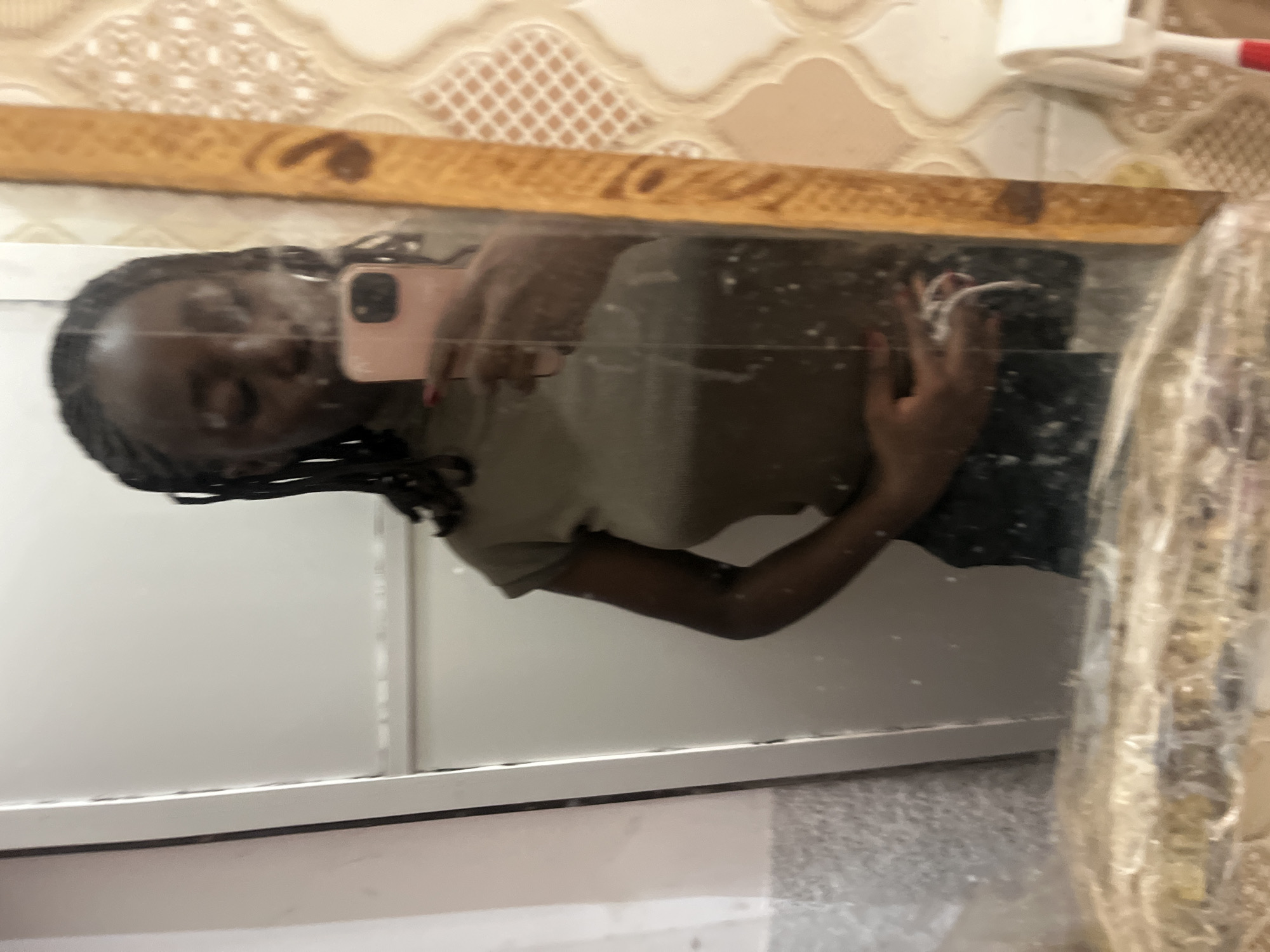 The image is a selfie taken in a bathroom in Abuja, Nigeria. A person with dark skin, hair styled in braids, is visible in the reflection of a mirror. The person is holding a phone with a pink case. The background includes patterned wall tiles and a sink area. A decorative object wrapped in plastic is also visible.