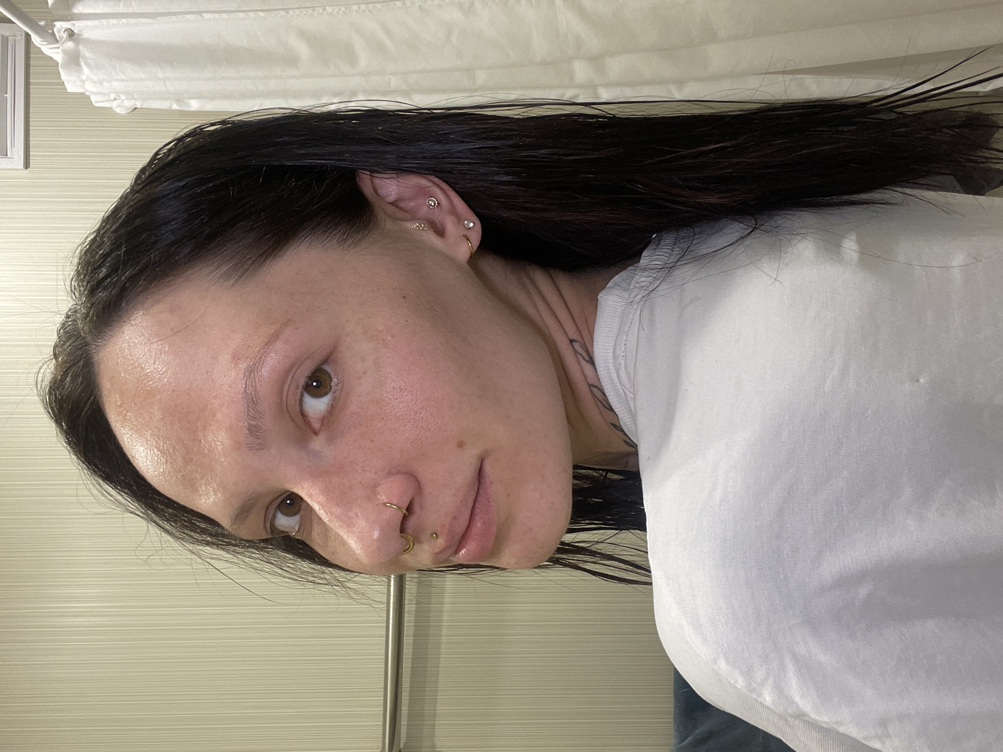 A medium shot shows a person with brown eyes and dark hair, wearing a white shirt. The person has multiple piercings in their left ear, including helix, lobe, and cartilage piercings. There are also piercings in the nose and lip. The hair appears wet and sticks to the forehead and side of the face. The background consists of a light-colored wall with subtle horizontal lines, and a white curtain or sheet hanging in the upper portion of the frame. The image does not contain enough information to confirm the location in Izhevsk, Russia.