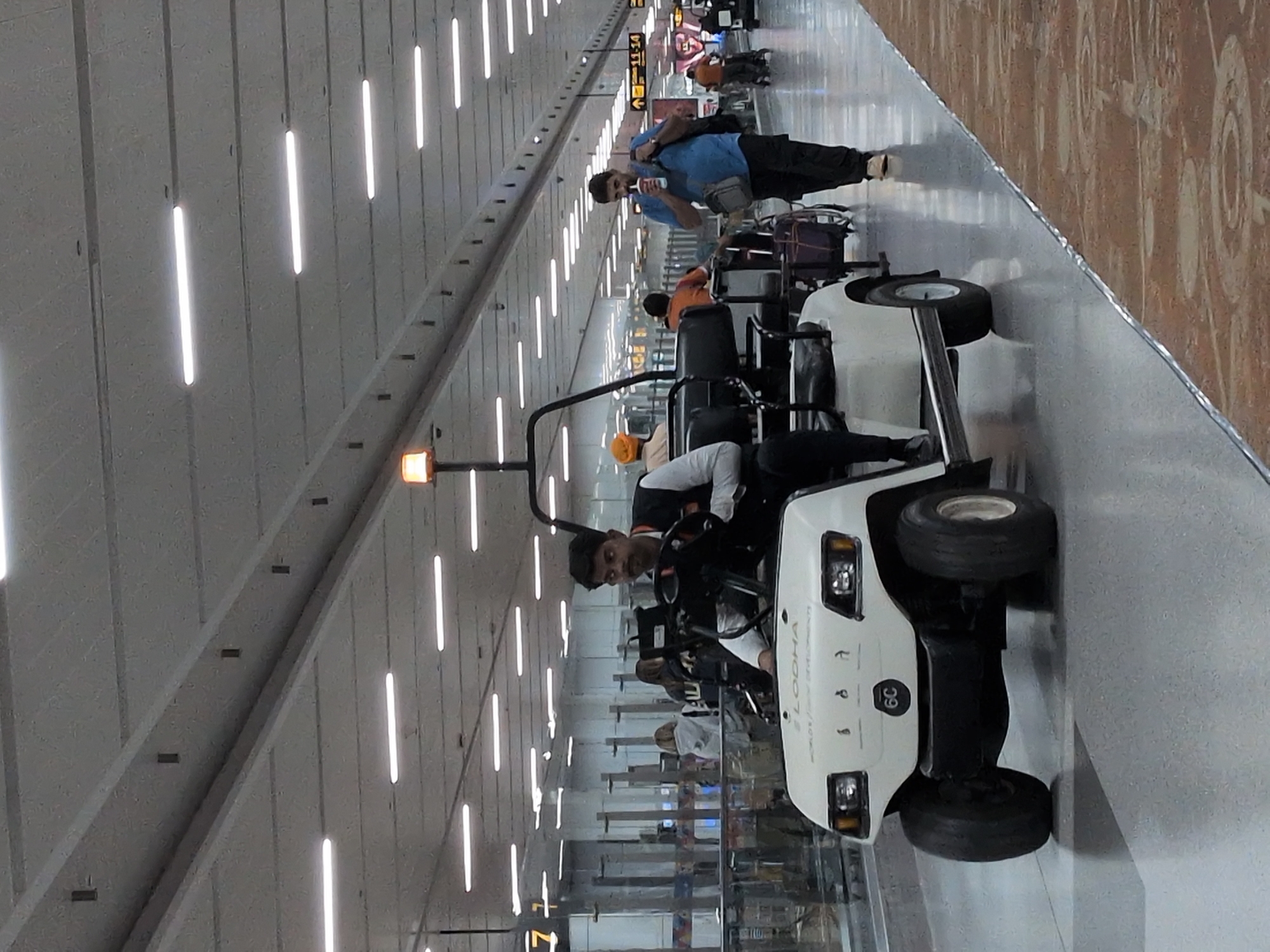 The image depicts an interior view of Indira Gandhi International Airport in New Delhi, India. In the foreground, a white electric golf cart, branded with "LODHA WORLD'S LARGEST DEVELOPMENTS" and "6C", is stationary. A male driver, wearing a black vest over a white shirt, is seated in the driver's seat. The ceiling features a grid of fluorescent lights. In the mid-ground and background, several individuals are visible walking in a hallway. One man in a blue shirt is seen drinking from a cup while walking. Another man in an orange turban is seated in the background. The overall environment appears to be a busy airport terminal.