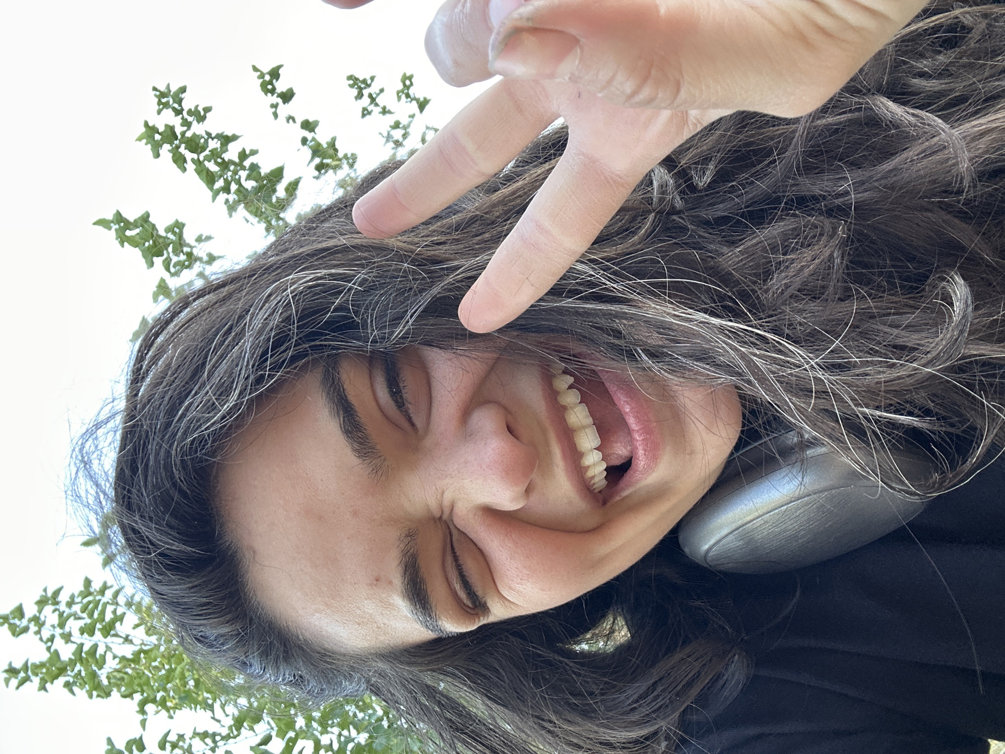 A young woman with dark, wavy hair and streaks of gray is pictured from the chest up, looking towards the camera with a wide smile and one eye winking. She is wearing a black t-shirt and headphones around her neck. Her right hand is raised, forming a peace sign. She is outdoors, with green foliage and a bright sky visible in the background. The image was taken at a low angle, making it appear as though the viewer is looking up at her. The scene is likely in Malevo, Bulgaria, given the outdoor setting and common foliage.