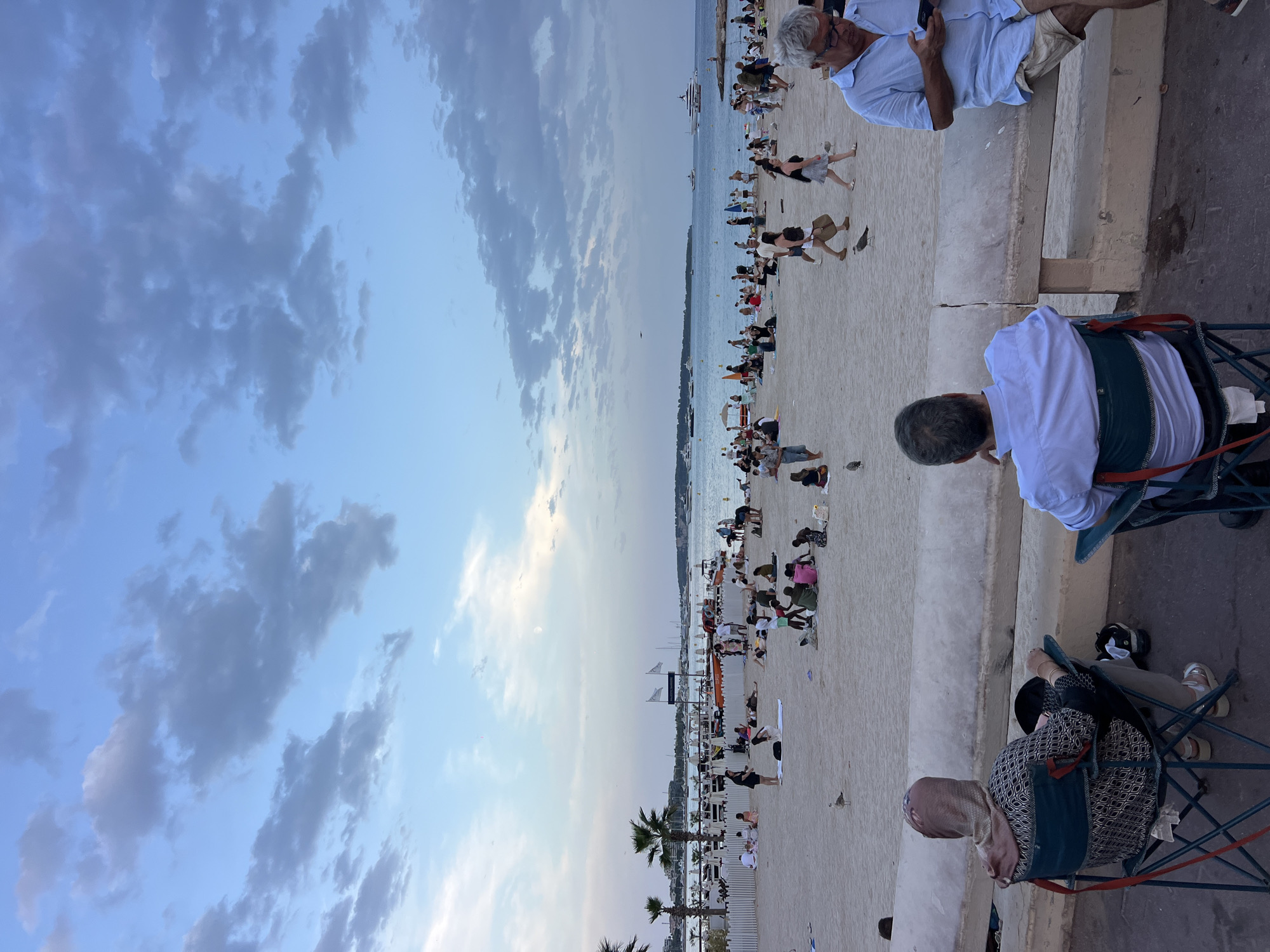 The image depicts a crowded beach scene in Cannes, France, likely during the late afternoon or early evening, given the soft lighting and cloudy sky. In the foreground, two individuals are seated on folding chairs, facing away from the camera and towards the beach. The person on the left wears a headscarf and a patterned dress, while the person on the right wears a light blue shirt and shorts.
The beach itself is filled with numerous people engaging in various activities. Some are lying down on towels, others are sitting in groups, and some are walking along the sand. There are beach umbrellas scattered throughout, suggesting a recreational area. In the background, a white fence separates the beach from a promenade or walkway, and beyond that, palm trees and buildings line the shore.
The sea is visible in the distance, with several boats and yachts anchored or cruising on the water. The sky is a mix of blue and grey clouds, with a soft glow indicating either sunrise or sunset. The overall atmosphere is one of leisure and public gathering, typical of a popular coastal resort town.