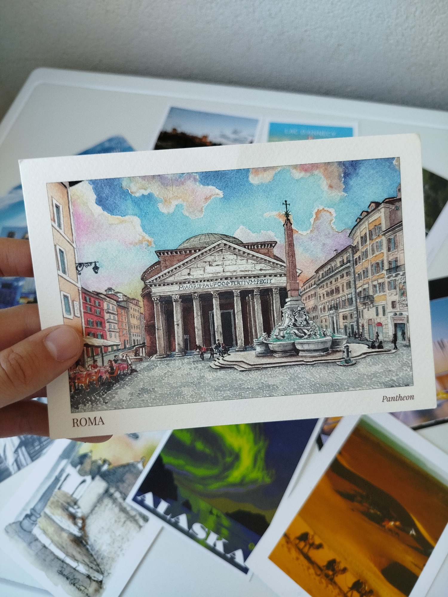 The image shows a postcard depicting a watercolor painting of the Pantheon in Rome, Italy. The postcard is held by a person. The painting shows the Pantheon, a fountain with an obelisk, surrounding buildings, and a cloudy sky. The word "ROMA" is printed on the bottom left of the postcard, and "Pantheon" on the bottom right. The postcard is surrounded by other postcards.