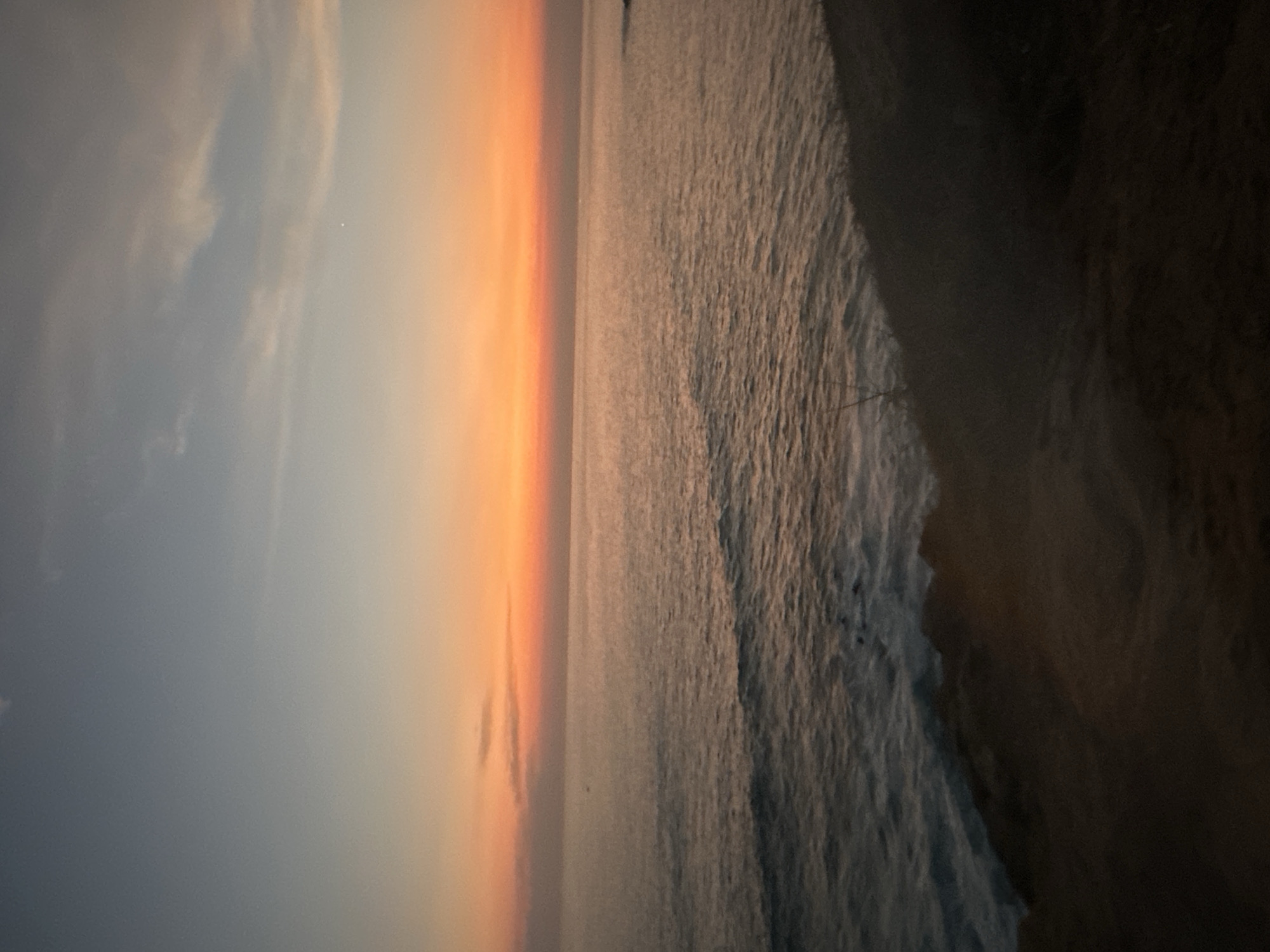 The image is a sideways view of the beach in Puerto Escondido, Mexico, possibly at sunrise or sunset. The sky is a gradient of grey and orange, meeting the ocean in a bright, diffused horizon. The ocean appears turbulent, with visible waves approaching the shore. The shoreline is dark and undefined, obscuring specific details.