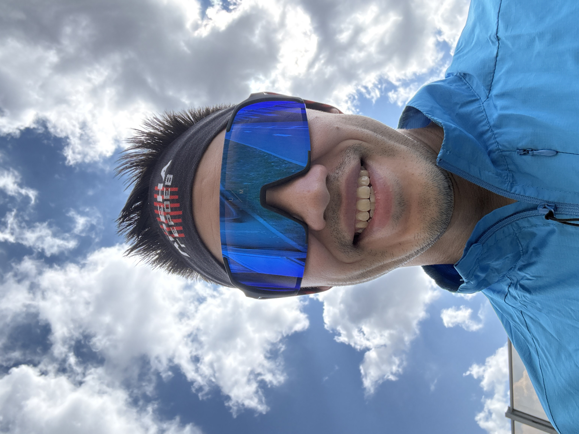 An eye-level or slightly low-angle close-up captures a person looking directly upwards at the camera, against a bright blue sky filled with scattered white cumulus clouds. The subject, likely male, is smiling widely, revealing teeth, and has short, spiky dark hair and light stubble on their face. They are wearing a vibrant blue athletic jacket with a visible zipper and a dark-colored headband that features red and white striped patterns and the partial text "HAB".
Prominently featured are large, wraparound athletic sunglasses with intensely reflective blue lenses. The reflection in the lenses appears to show a road or track extending into the distance, possibly indicating an outdoor sporting or travel activity. The letters "UM" are faintly visible on the left lens. The overall impression is one of joy or exhilaration, perhaps after or during an outdoor event.
The bright daylight suggests it is daytime. While the primary background is the sky, one of the cropped images faintly reveals what looks like a modern building structure with glass and metal elements in the lower right corner, hinting at an urban or developed outdoor setting, consistent with the context of Kazan, Russia. The person's active wear and cheerful expression suggest a moment of enjoyment during a physical activity in good weather.