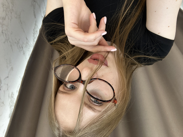 The image presents a close-up, slightly off-center portrait of a young woman situated indoors, reportedly in the city of Chaikovsky, Russia. The subject has long, straight, light-colored hair that partially obscures the right side of her face and falls over her shoulders. She wears tortoise-shell framed eyeglasses, positioned somewhat askew with the right lens appearing lower than the left, through which her blue eyes are directed forward. Her lips are painted with pink lipstick and are slightly parted. She is dressed in a black short-sleeved t-shirt. Her right hand, exhibiting light pink manicured fingernails, is raised with the index finger gently touching the area near her lower lip or chin. Visible moles are present on her right forearm. The background is divided, showing a light brown or beige fabric curtain on the left side and a white and gray marble-patterned surface on the right, indicative of an interior setting.