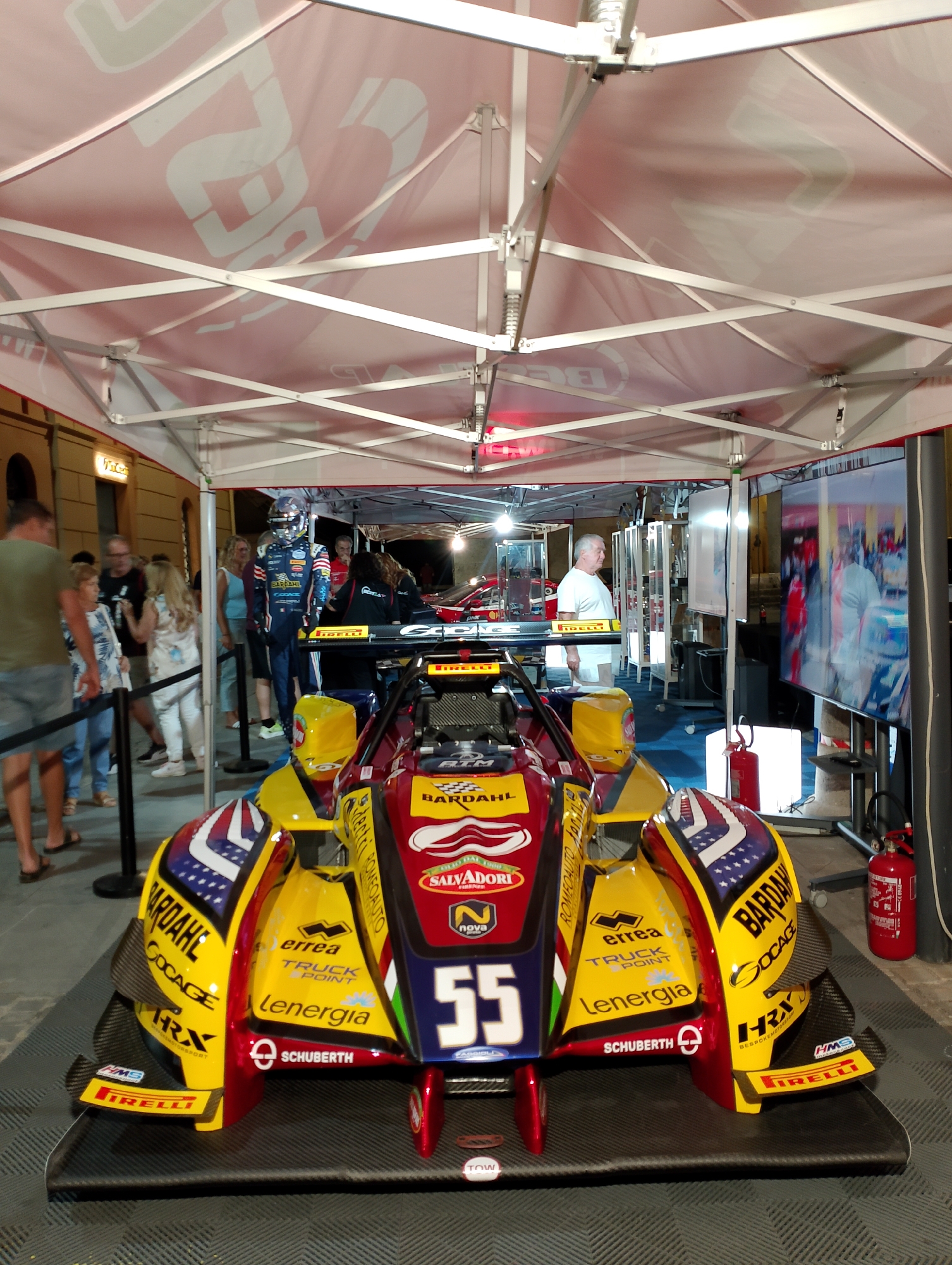 The image shows a race car displayed under a red tent. The car is predominantly yellow and red with various sponsor logos, including "ERREA", "LENERGIA", and "SCHUBERTH". A mannequin dressed in a racing suit is standing to the left of the car. Several people are visible in the background, some of whom appear to be spectators. A large screen displaying footage of a race is on the right side of the image. A fire extinguisher is mounted on a stand to the right of the car. The scene appears to be set up outdoors, possibly at an event or exhibition. The presence of the "Salvadore" logo on the car suggests a connection to Rieti, Italy, as Salvadori is a known brand from that region.