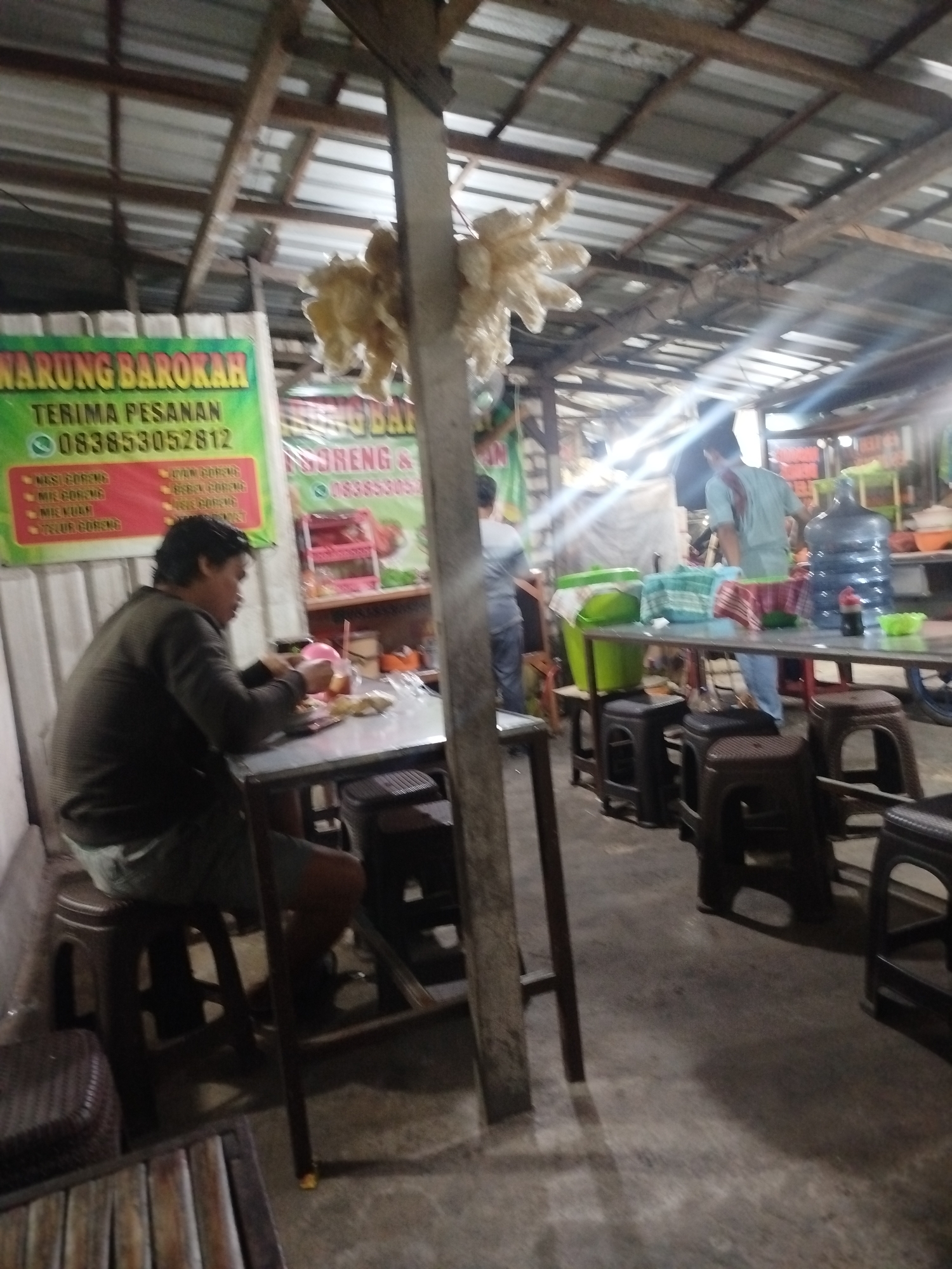 This is a photograph of an outdoor food stall in Tonjung, Indonesia. The stall appears to be a casual dining establishment, likely a warung, with a tin roof supported by wooden beams. A banner on the left side of the image advertises "WARUNG BAROKAH" and lists food items and a phone number. In the foreground, a person is seated at a metal table, eating. They are wearing a dark shirt and shorts. To the right, in the background, other individuals are visible, including one wearing a light blue shirt and standing near large water jugs. Various plastic containers, likely for food storage or serving, are also present on tables and shelves. The overall environment is rustic and functional, typical of informal food vendors.
