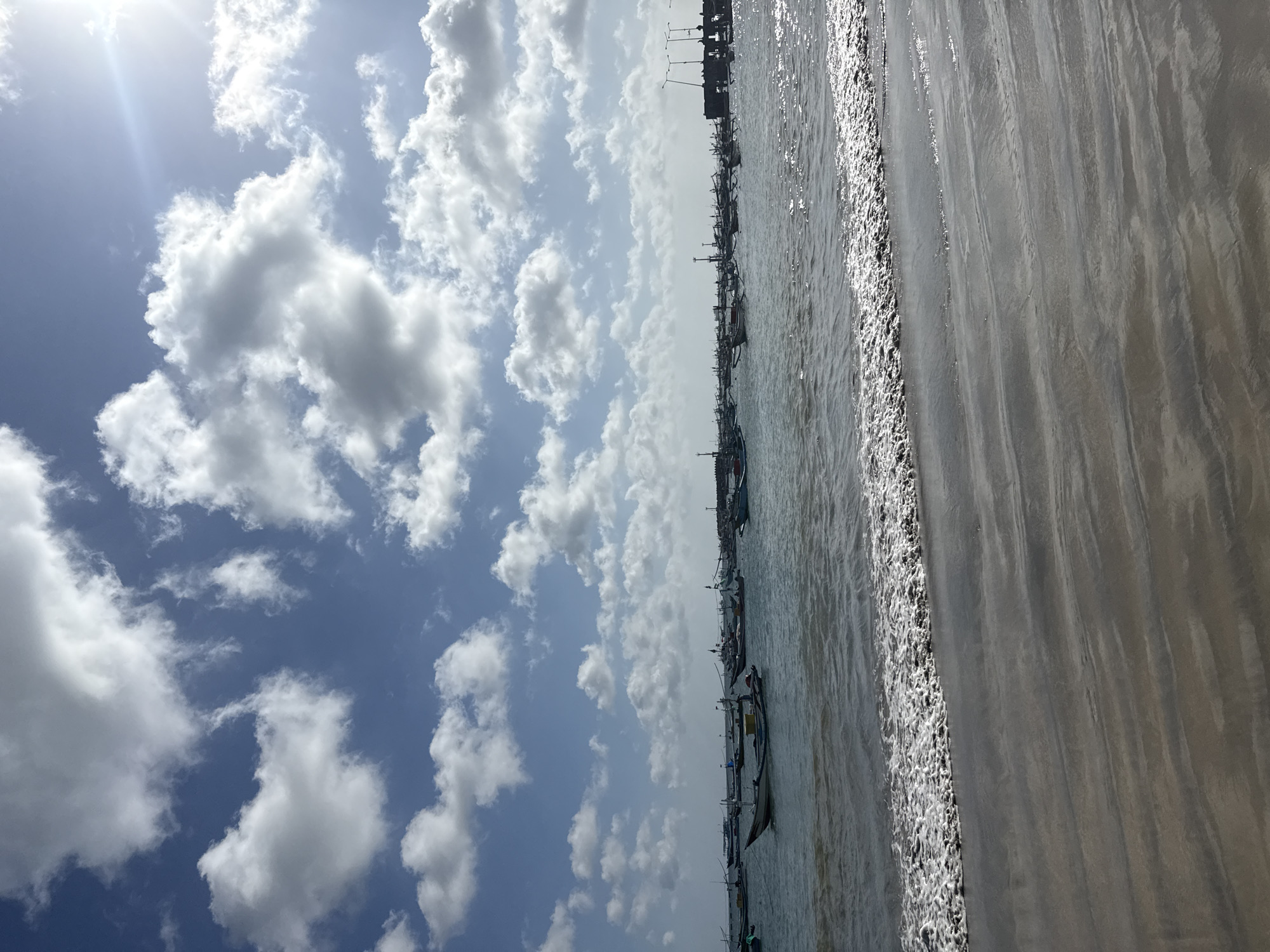The image shows a beach scene in Kuta, Indonesia. The sky is blue with white cumulus clouds. The sun is shining brightly. The beach has wet sand and the ocean is reflecting the sunlight. Several fishing boats are anchored in the water. The image is oriented in portrait mode.