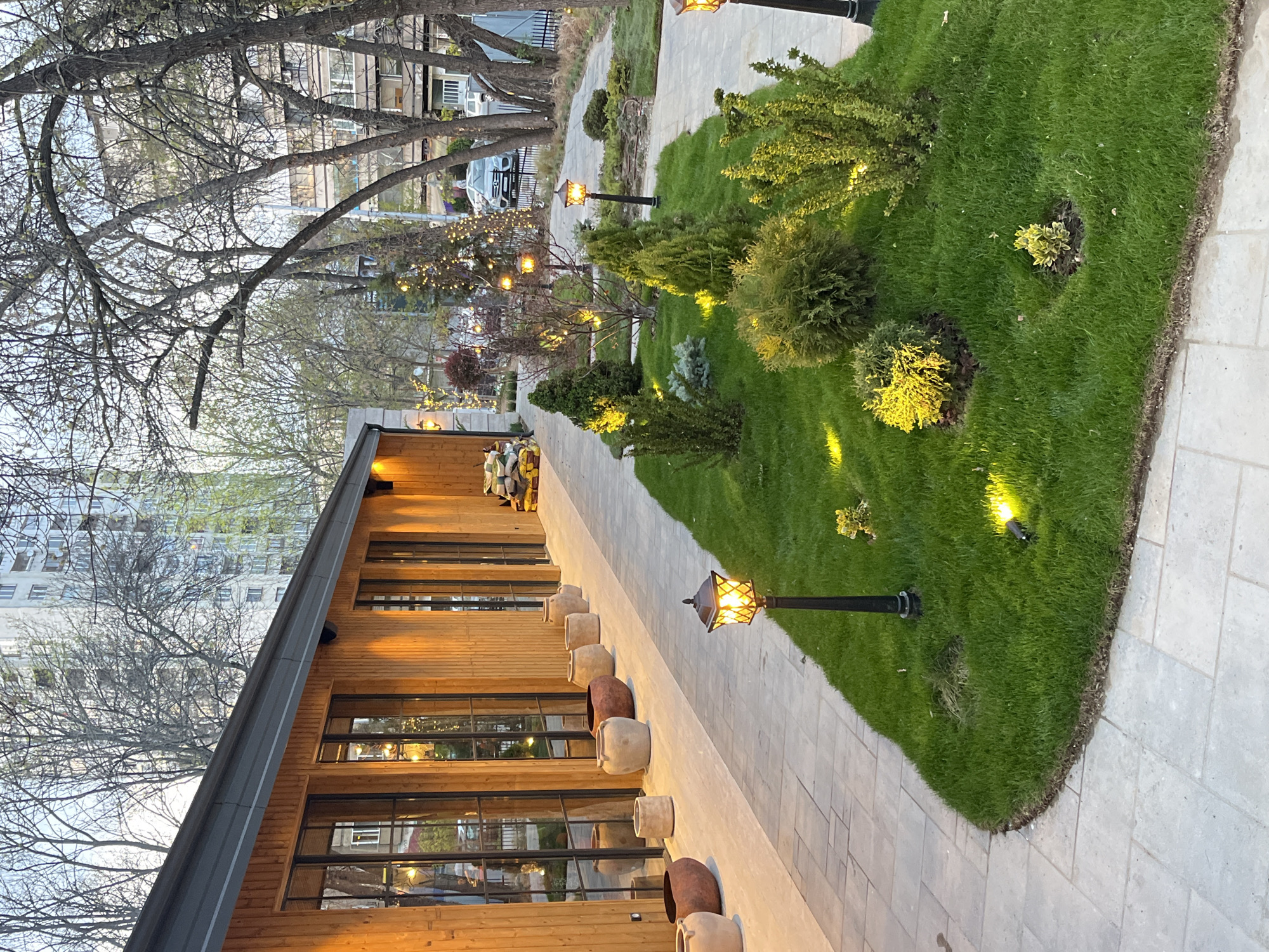 This image captures an inviting outdoor scene, likely a restaurant or cafe patio, during dusk or early evening in Sofia, Bulgaria. The artificial lights are on, casting a warm glow across the area.
On the left side, a modern building features warm wooden siding and large, dark-framed windows that reflect the ambient light. Through these windows, the interior lights are visible, along with faint silhouettes of people, suggesting patrons dining or socializing inside. Along the base of the building, a line of decorative ceramic pots in varying earthy tones and sizes are placed. The roofline of the building is visible towards the top left, featuring a distinct grey gutter.
To the right of the building, a light-colored paved walkway extends into the distance, running parallel to the structure. Adjacent to the path is a lush, well-maintained green lawn interspersed with various small, neatly trimmed shrubs and plants. Several ground-level spotlights illuminate these plants, creating bright yellow highlights. Black streetlights with ornate, amber-glowing lanterns are positioned at intervals along the paved path, providing additional illumination. Further into the garden, smaller trees are decorated with strings of warm fairy lights, enhancing the cozy atmosphere.
In the background, bare-branched trees suggest an early spring or late autumn setting, partially obscuring larger, light-colored multi-story buildings and a white SUV parked behind a fence. The sky still holds some light, indicating that the sun has recently set. The overall mood conveyed is peaceful, welcoming, and pleasant. There is no visible text in the image.