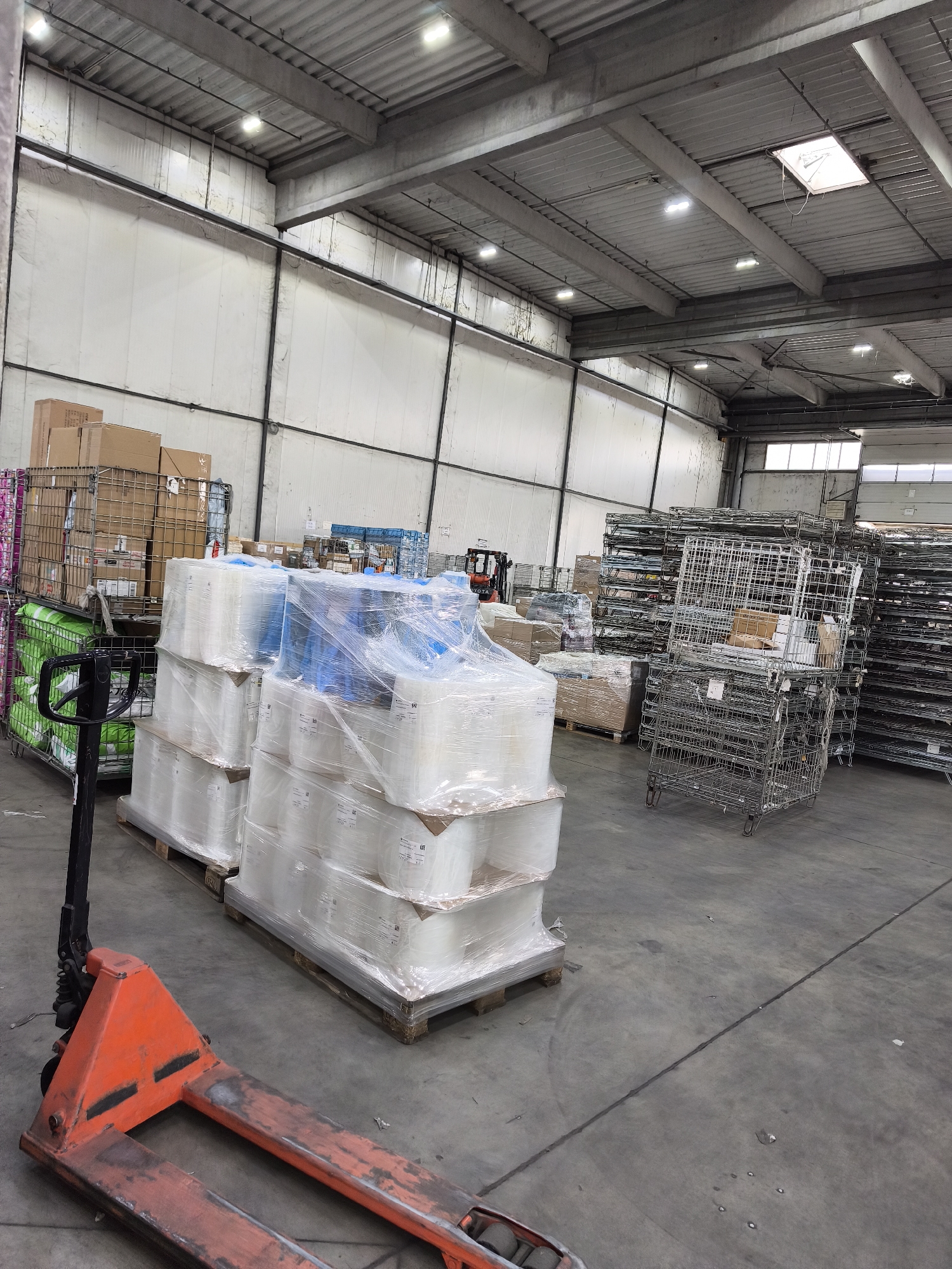 The image presents the interior of a large industrial warehouse or storage facility, likely located in Burgas, Bulgaria, given the context. The scene is well-lit by numerous rectangular LED light fixtures embedded in the high, corrugated metal ceiling, which also features several skylights.
In the immediate foreground, an orange and black manual pallet jack rests on a grey concrete floor, which shows signs of use and minor debris. To the left, two pallets are stacked with white, bucket-like containers or packages, tightly wrapped in clear plastic film. Some blue items are visible beneath the plastic on the uppermost pallet. Adjacent to these, a metal cage trolley contains vibrant green bags, topped with cardboard boxes.
The mid-ground on the right is dominated by stacks of empty, foldable wire mesh containers, some equipped with wheels, suggesting they are used for transport or storage of goods. In the far background on the right, a red forklift, identifiable by the visible "TOYOTA" logo, is partially obscured by other stored items and equipment. The walls of the warehouse are composed of tall, white, insulated-looking panels with black support beams running horizontally and vertically.
No active event or human activity is depicted; the scene captures a static moment within what appears to be an organized industrial storage or staging area. The time of day appears to be daytime, aided by natural light from the skylights. Visible text includes the "TOYOTA" logo on the forklift and small, unreadable product labels on the white wrapped containers, faintly showing numbers like "132" and "190" and partial dates such as "01/202_".