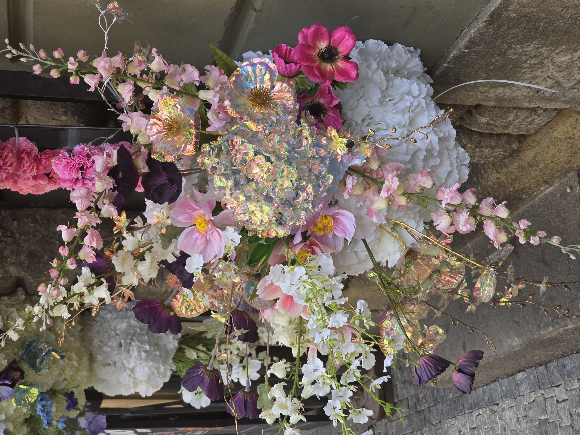 A large, elaborate floral arrangement dominates the scene, mounted against a textured stone wall. The display consists of a diverse collection of artificial flowers and iridescent, transparent decorative elements. Predominant colors include white, various shades of pink (from pale pastel to vibrant fuchsia), deep purple, and scattered hints of light blue.
Specific floral forms include dense, spherical clusters of white petals (resembling hydrangeas or carnations) at the lower left and right. Numerous delicate, bell-shaped flowers in white and pale pink are arranged on arching stems. Brighter pink, flat-petaled flowers with yellow centers and fuchsia anemone-like flowers with dark centers are interspersed throughout. Deep purple, drooping bell-shaped flowers are visible at the lower left.
The iridescent components feature larger, stylized floral shapes with clear centers and numerous smaller, geometric pieces clustered together, reflecting light with a rainbow sheen. Thin, metallic gold-colored decorative branches are interwoven within the arrangement.
The background consists of a rough, light brown stone or concrete wall on the right and behind the flowers. To the far left, a dark, vertical structural element, possibly a metal frame, is partially visible. At the bottom left, grey cobblestone paving and a stone curb are present, indicating an outdoor, urban setting. No people are visible in the image.