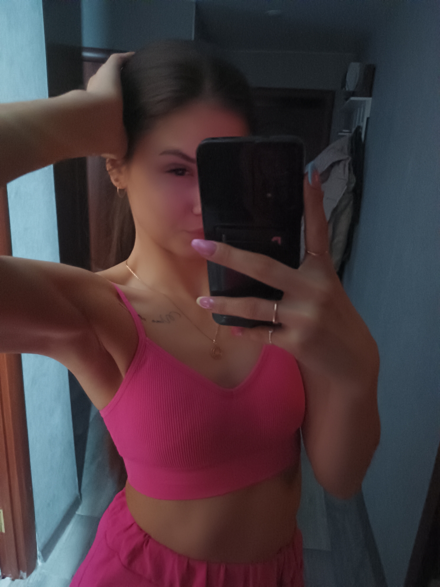 A young woman is taking a selfie in what appears to be an indoor setting, possibly a bedroom or apartment. She is wearing a bright pink two-piece outfit consisting of a crop top and shorts. Her hair is dark and pulled back, and she is wearing jewelry, including earrings and a necklace. She is holding a smartphone up to her face, obscuring part of her face with the phone. The background includes a door, a wall painted blue, and possibly a coat rack with clothes hanging on it. The location is unknown.