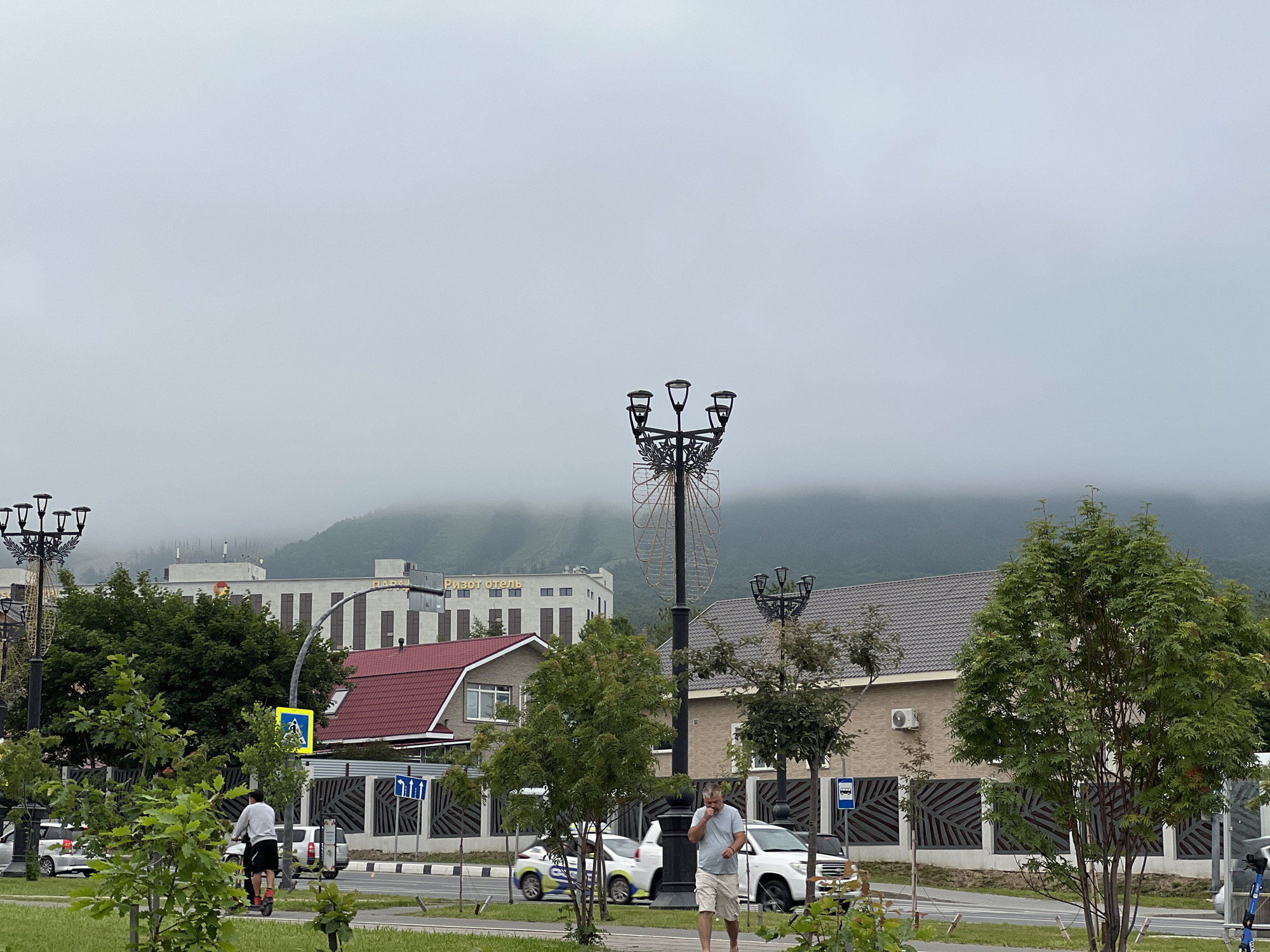 A street scene in Yuzhno-Sakhalinsk, Russia, on an overcast day shows a man on an electric scooter riding on a sidewalk. In the background, there are buildings, including one with a sign that reads "РИЗОТ ОТЕЛЬ" (Rizot Hotel), a pedestrian crossing, streetlights, trees, and cars. A second man is walking on the sidewalk, holding his phone to his ear. The environment appears to be a city park or pedestrian area adjacent to a road.