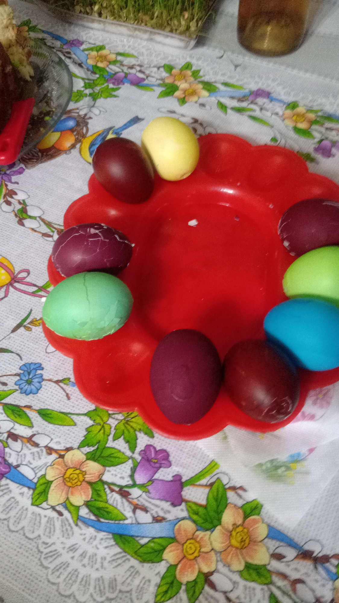 The image depicts a red, flower-shaped plastic plate filled with approximately nine colored Easter eggs in a loose ring around the plate's center. The egg colors are varied, including brown, yellow, purple, green, and blue. Some of the eggs show cracked shells. The plate sits on a patterned tablecloth featuring floral designs. In the background, there are blurry images of a cake, a small container of sprouting seeds, and a jar of liquid. There is no information to confirm or deny the location in the city of Yerevan, Armenia.