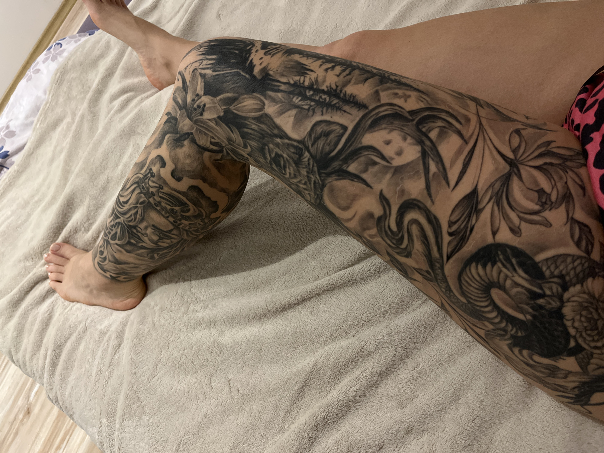 The image shows a person's legs resting on a beige blanket. The left leg, which is prominently displayed, is covered in a large, detailed black and grey tattoo featuring floral designs, a snake, and a landscape. The person's foot is visible at the bottom left of the image. The background consists of the blanket and a portion of a wooden floor in the lower left corner. The location is not discernible from this image.